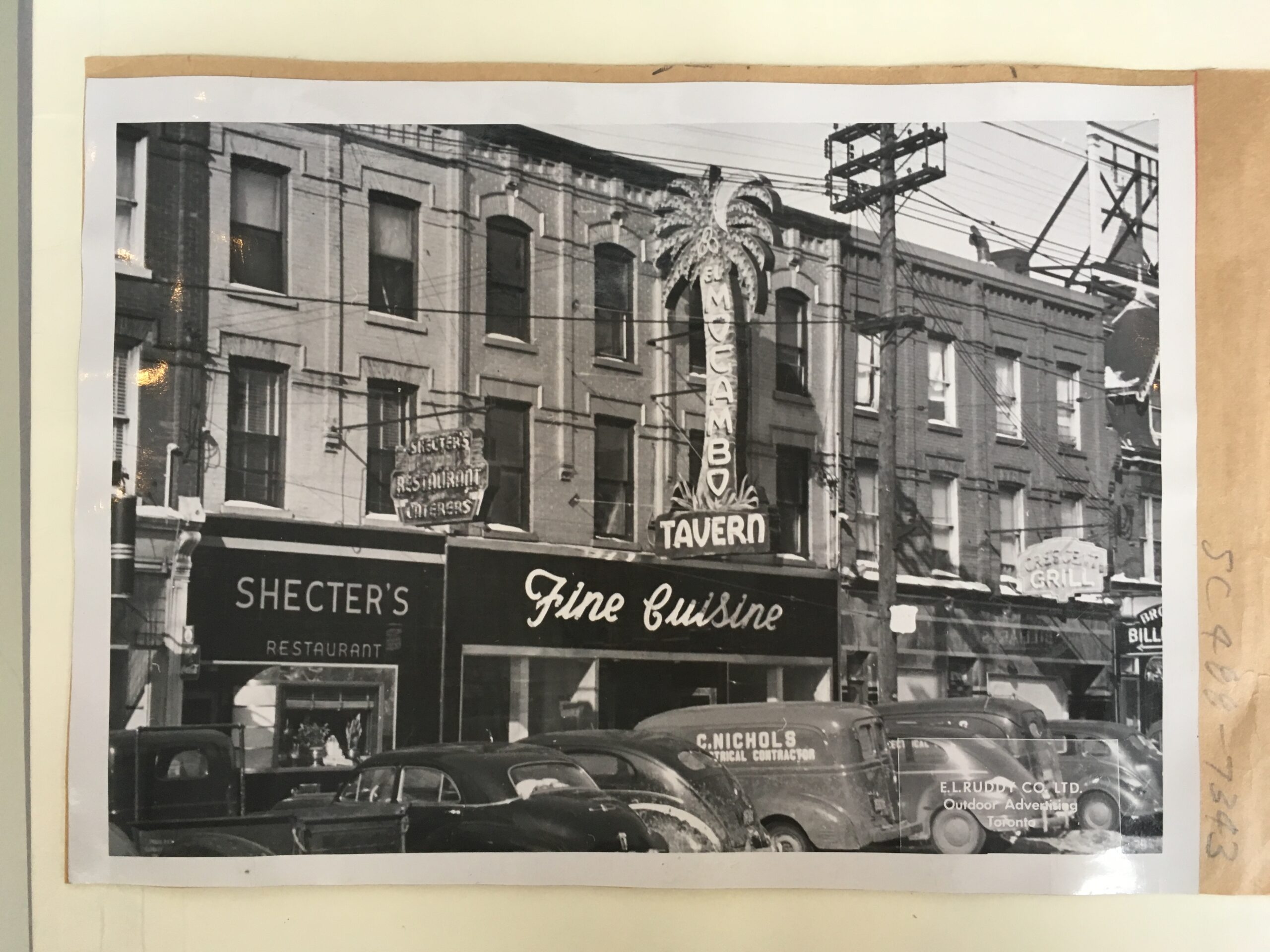 A black and white image showing a Toronto city block. On the right is a building featuring a neon sign in the shape of a palm tree that reads “El Mocambo Tavern”. Below reads “Fine Cuisine.”