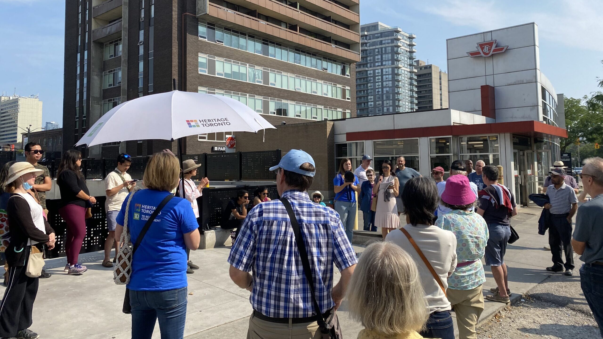 A large group of people gather on a paved sidewalk. In the foreground, a woman wearing a blue t-shirt holds a white umbrella. In the background, a woman in a pink dress speaks into a microphone. On the right, a symbol of the TTC can be seen atop a modern subway station.