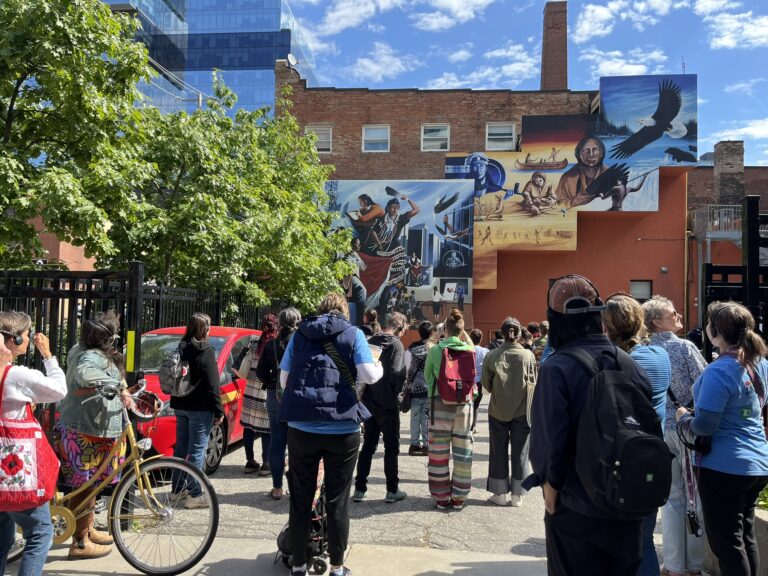A large group of people gather in front of a brown building with multiple murals on its outer walls. Pictured in the murals, among multiple aspects, are eagles, Indigenous characters, and river scenery.