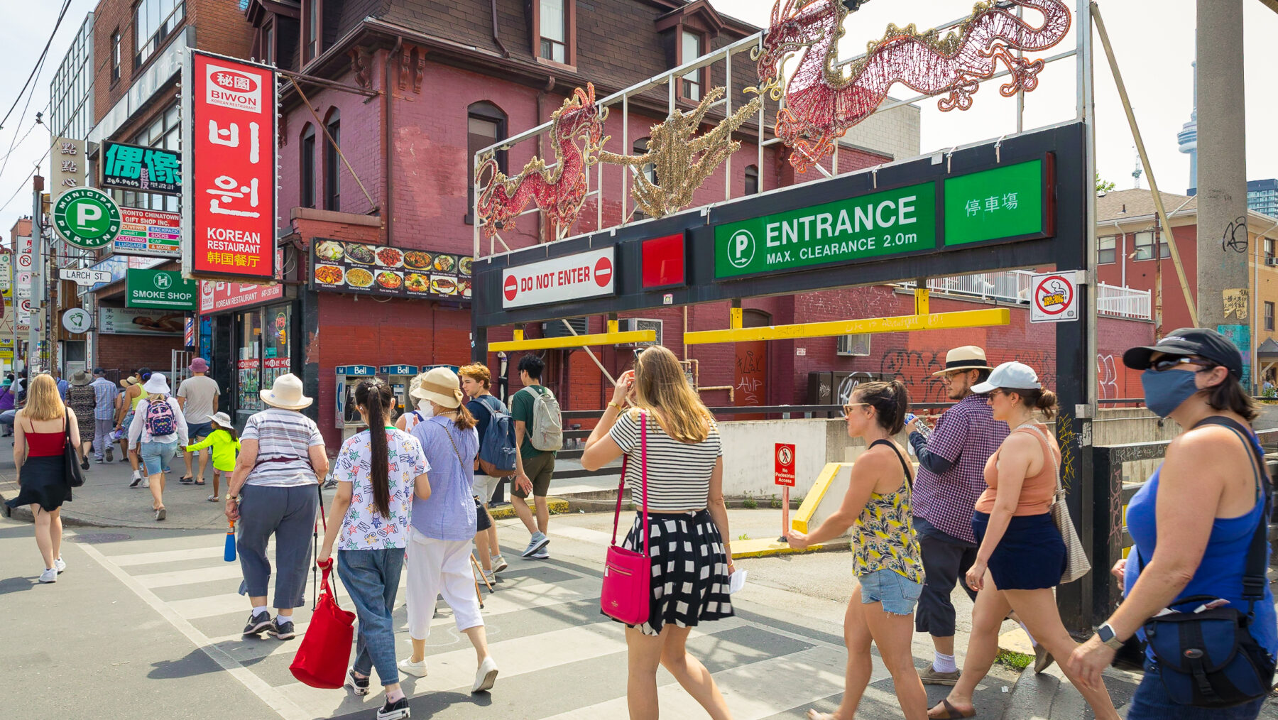 Line of people walk across a city street front. Signage features Chinese characters and parking lot entrance lighting in the shape of two large dragons.