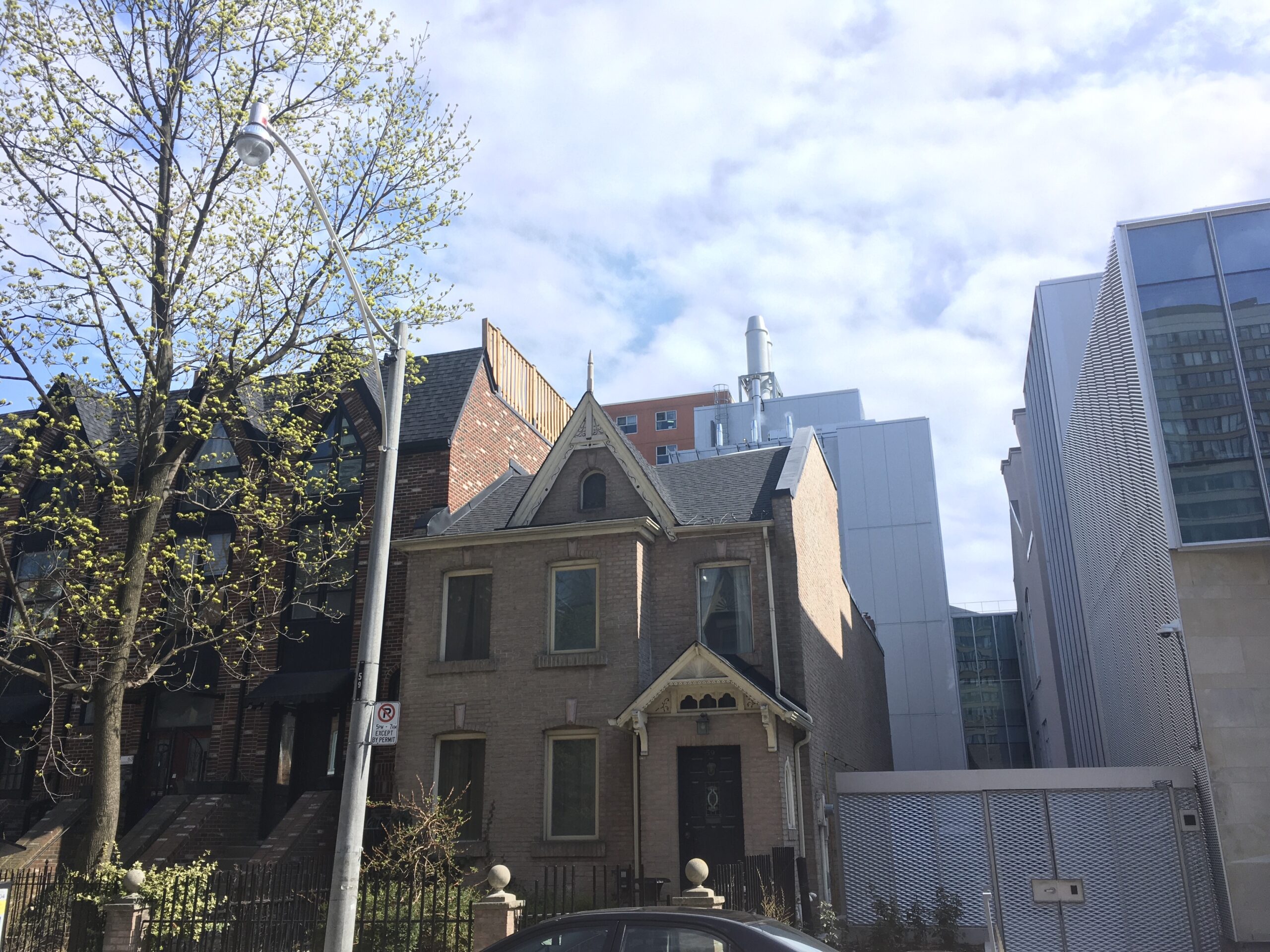 Colour photo of a light brick house. It is three storeys. The front of the house is surrounded by an iron gate fence. There is a street light in front of the house and to the left is a tree. Directly to the left of the house is a brick complex of semi-detached townhouses. Directly to the right is a metal gate and a modern style building. In the background are more buildings and a blue slightly cloudy sky.
