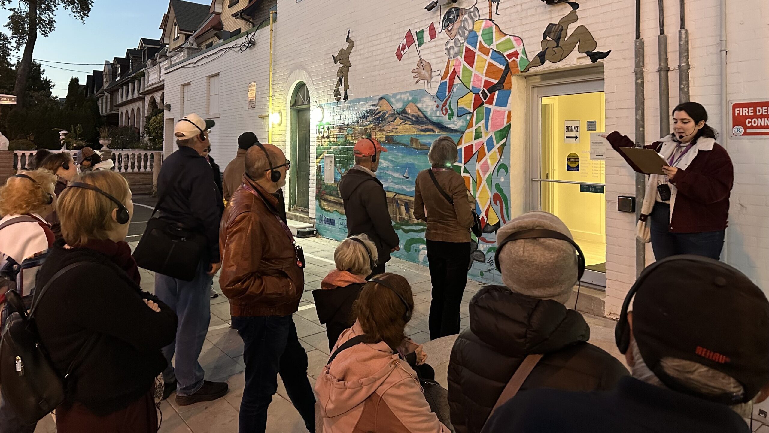 A group of people gather in front of a mural against a white-walled building. In front of them, a person with a headset microphone stands holding a clipboard and gestures to the mural.