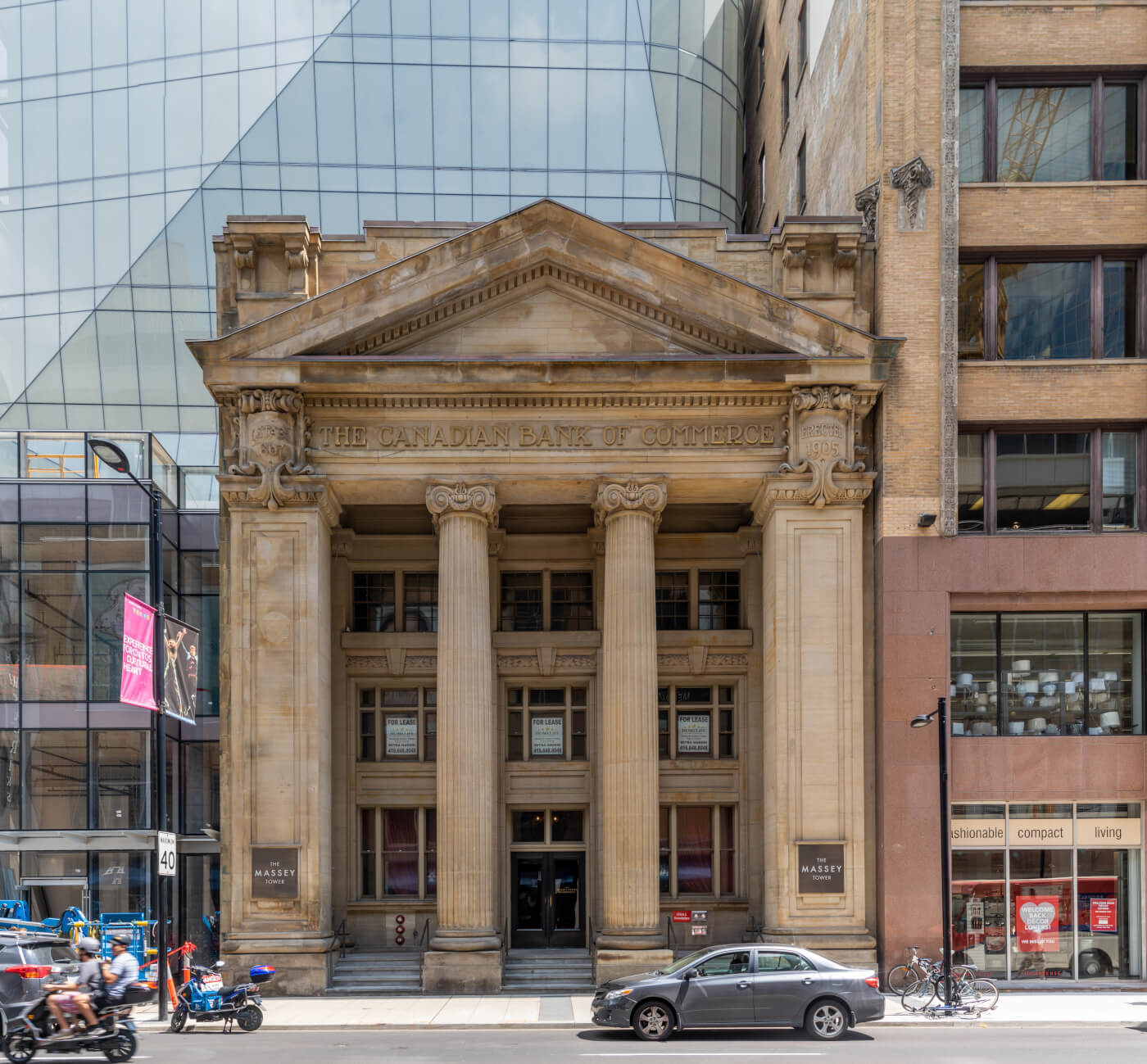 Photograph of a light coloured stone building set between modern buildings. There are two round pillars in the middle. The roof comes to a point with slightly marbled colour. Letting right below the roof reads "THE CANADIAN BANK OF COMMERCE". Below are large rectangular windows which extend to ground level. On either side of the stone building are brown plaques on the walls which read "THE / MASSEY / TOWER".