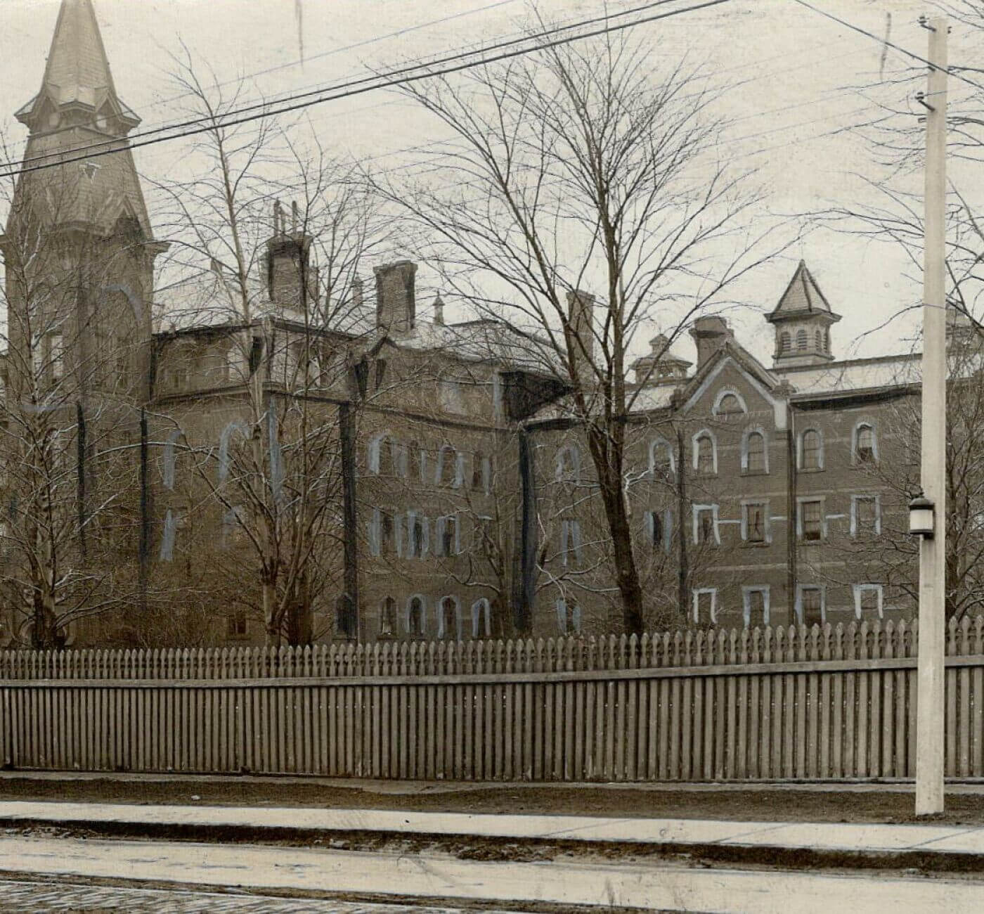 A black-and-white image of a large building complex. The buildings are made of brick and have many windows. There are also a number of towers with pointed roofs attached to the buildings. Around the complex is a wooden fence.