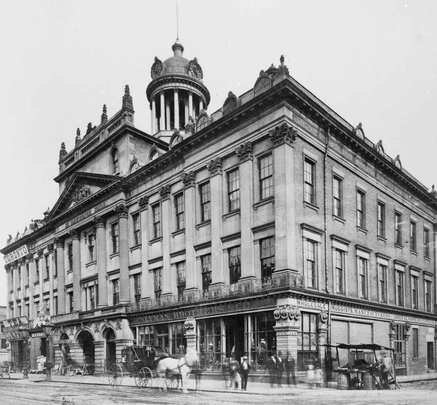 An archival photo shows St. Lawrence hall, a three story high large building with many windows and intricate stone detailing. A horse and buggy can see seen in front of the building along with the store front names "Lyman Brothers & Co, Druggists" as well as "Wholesale Druggists, Manufacturers, Linseed Oil, Paints, Dye-Stuffs".