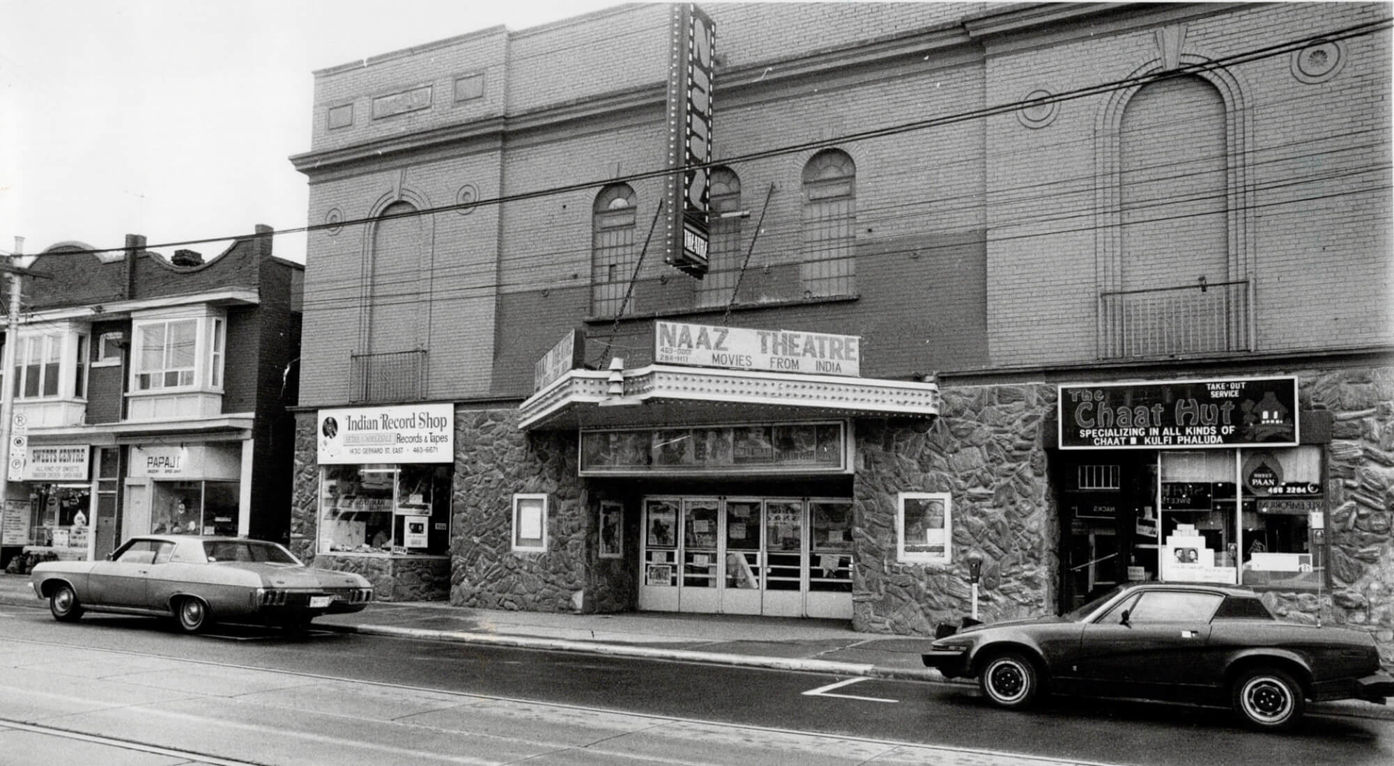 Black and white image of the front of Toronto's Naaz Cinema. The theatre is flanked by an Indian record shop and an Indian restaurant. There are 1980's cars parked in front of the theatre.