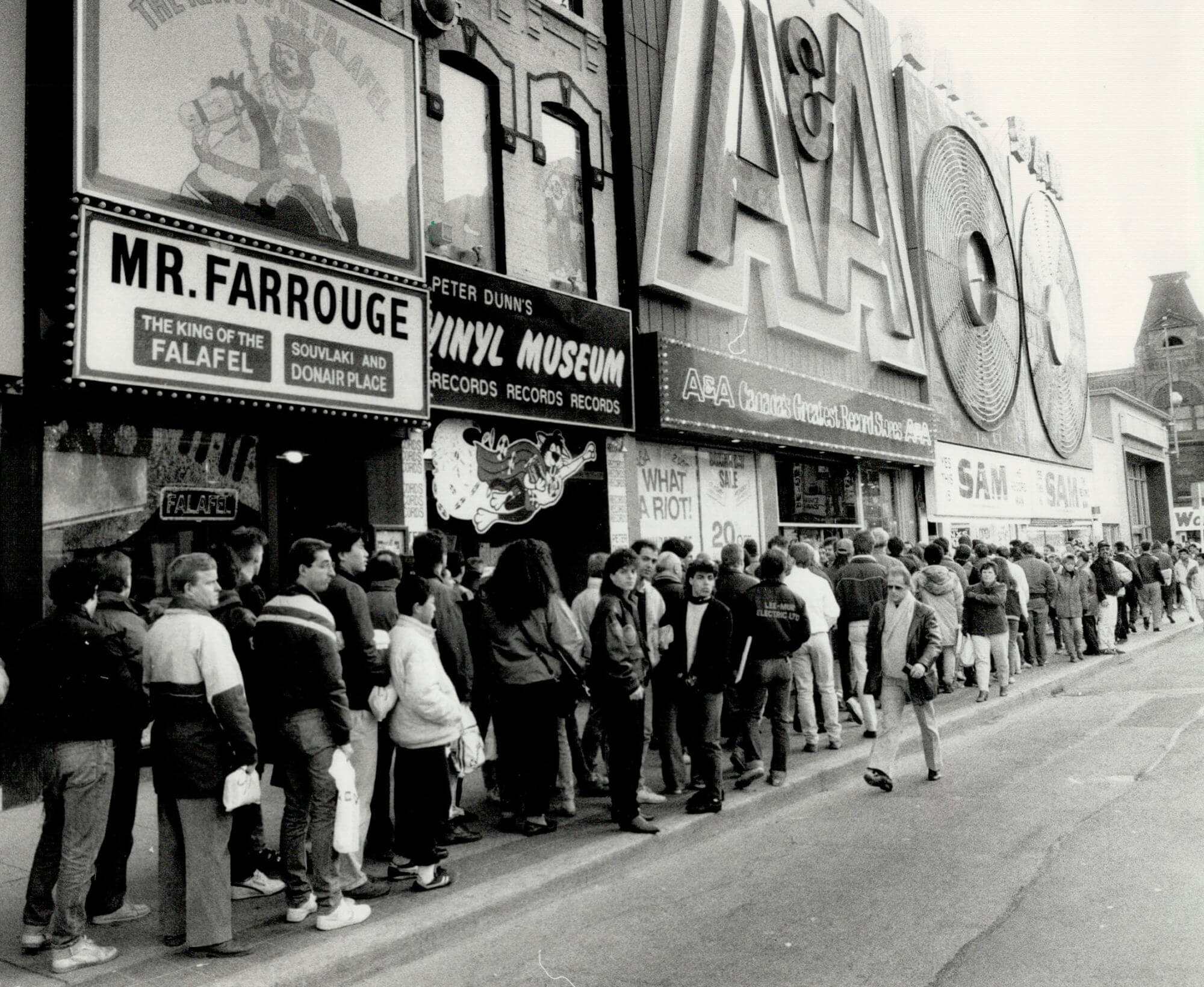 Black and white image of one side of Yonge Street. Sam the Record Man and A&A Record Stores are visible. There is a huge line up of people taking up the entire side walk. The Vinyl Musuem and a falafel restaurant are also visible.