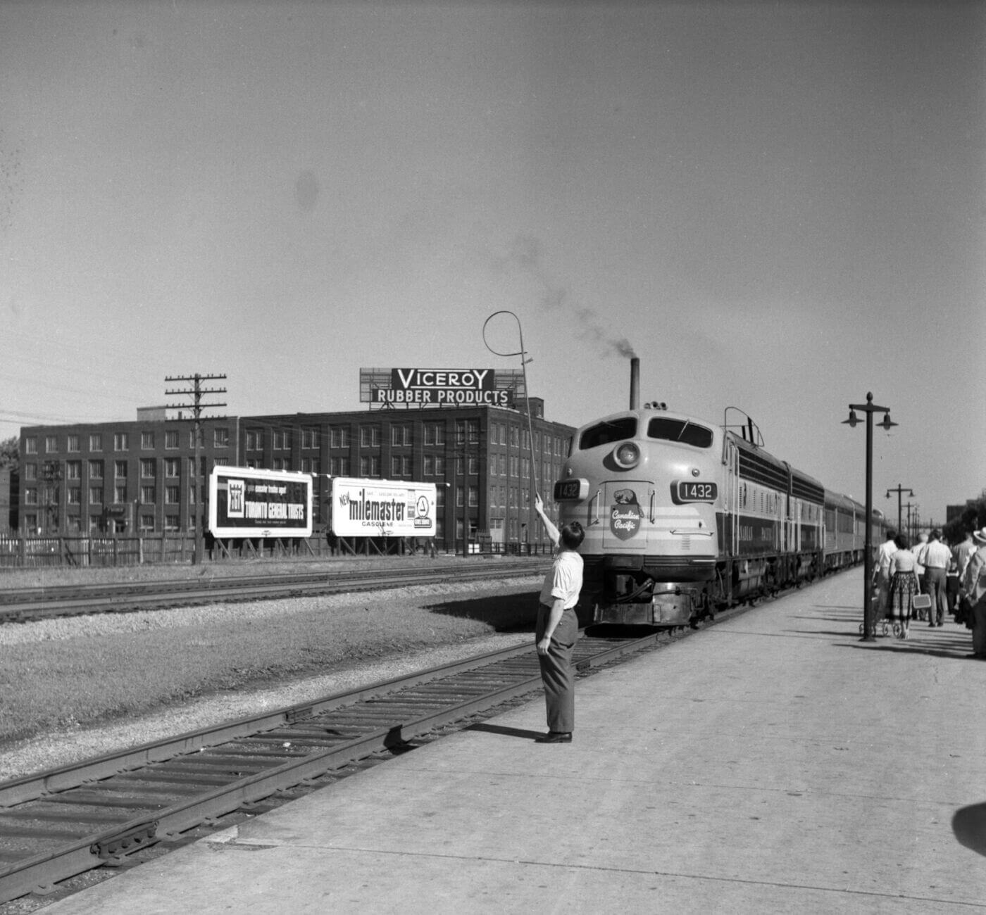 Black and white photo of a man in 50s clothing standing on a train platform waving at the approaching train. In the background is a building with a large sign that reads, "Viceroy rubber products". Two billboards it along side the opposite side of the tracks with advertisements on them, and a group of people walk away from the camera on the train platform in the corner of the photo.