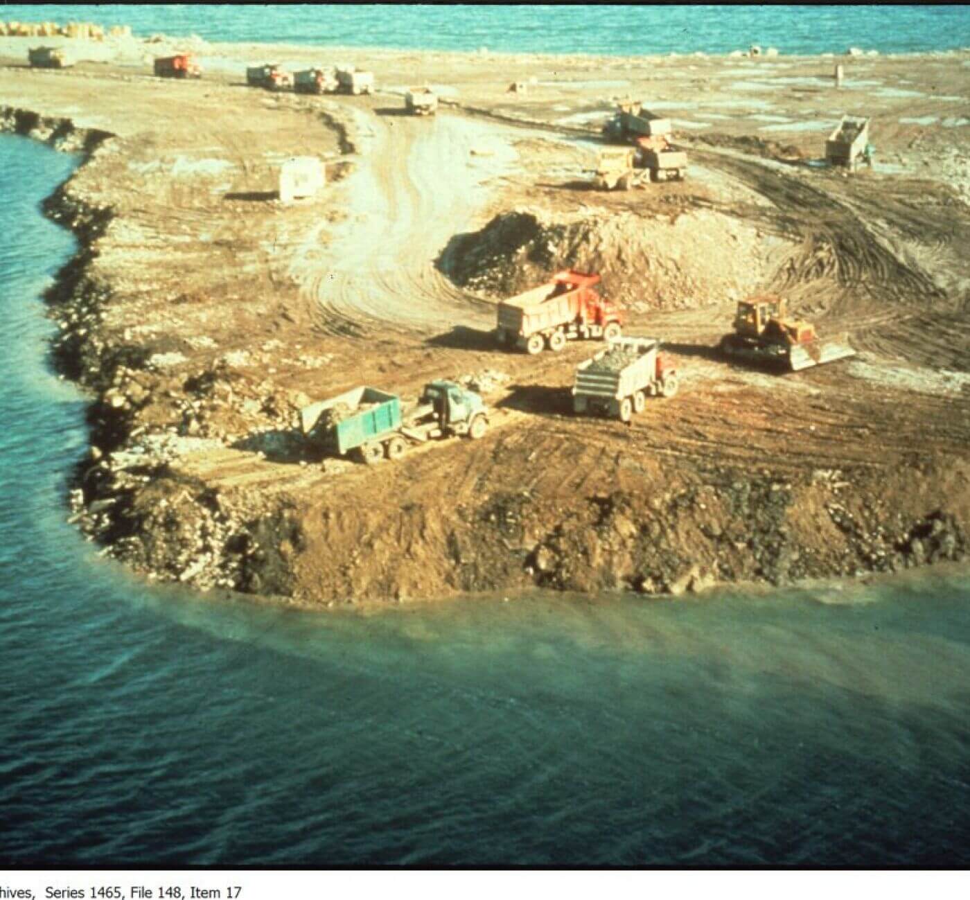 Colour photograph looking down on a rocky, brown strip of land, which takes up most of the image. Surrounding it is dark blue water, except for a strip of land at the top-left of the picture. No sky is visible. A dirt trail that leads from the lop-left stip down to the centre of the landmass. A mound of earth is situated there. Several large dump trucks move to and from this mound, taller than the three dump trucks closest to it. Another yellow construction vehicle is nearby. One of the dump trucks, painted blue, appears to be backing up and offloading soil or gravel in its trailer close to the water's edge. There are many ripples in the water, and some brown earth is visible by the shore under the water.