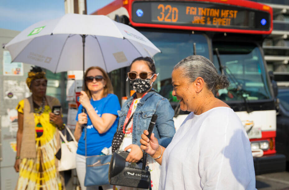 A photo of a group of four people standing on a sidewalk. There is a large red and white bus behind the group. Front left to right: person 1 in a yellow dress and headwrap, person 2 is in a blue t-shirt and holding a large white umbrella, person 3 is wearing a blue denim jacket and a face mask, person 4 is wearing a white t-shirt and speaking into a microphone.