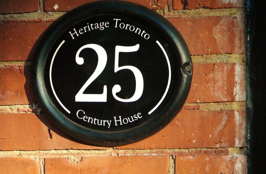 Image of a plaque mounted on a red brick wall. The plaque is oval in shape, with a black background and text that reads Heritage Toronto - 25 - Century House.