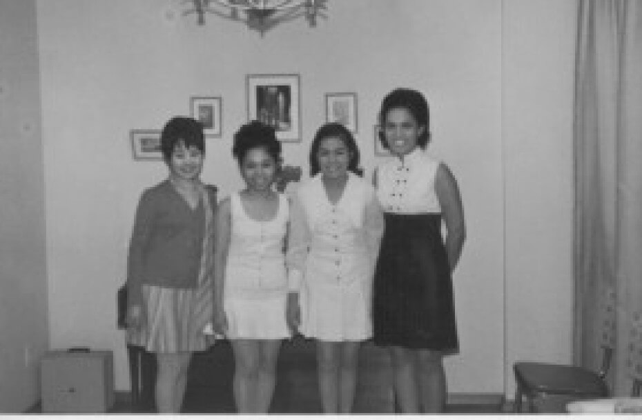 A black and white image of a group of four Filipino women standing together smiling and posing for the photograph in what looks to be a living room.