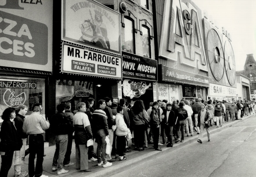 Black and white image of one side of Yonge Street. Sam the Record Man and A&A Record Stores are visible. There is a huge line up of people taking up the entire side walk. The Vinyl Musuem and a falafel restaurant are also visible.