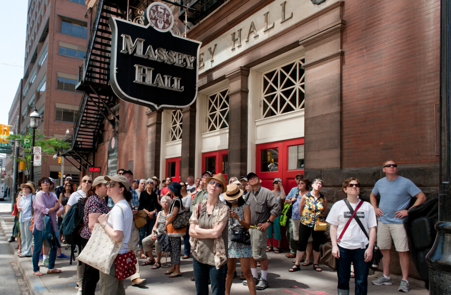 Image of a crowd, lightly attired, standing in front of the main red doors of Massey Hall looking up and away from the building, most face north. The main entrance is displayed prominently in the background with the Massey Hall neon sign in the upper centre of the photograph.