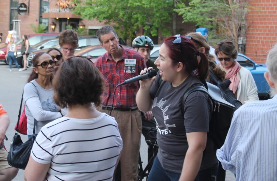 Image of a large group of people gathered around a tour leader wearing a volunteer shirt and speaking into a microphone. A parking lot is visible behind them.