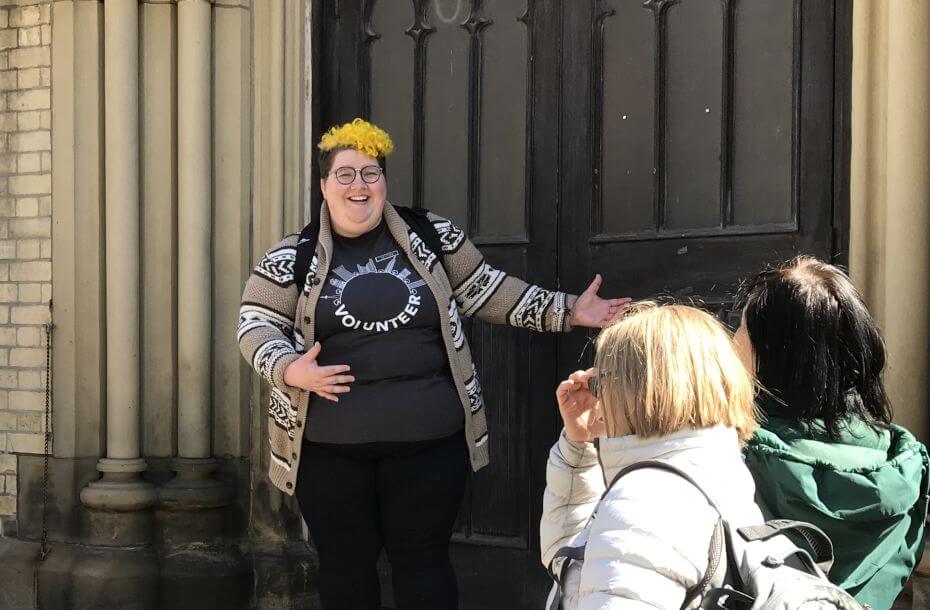 Image of young person standing on steps in front of side entrance of St James Cathedral. Participants of walking tour are gathered around them.