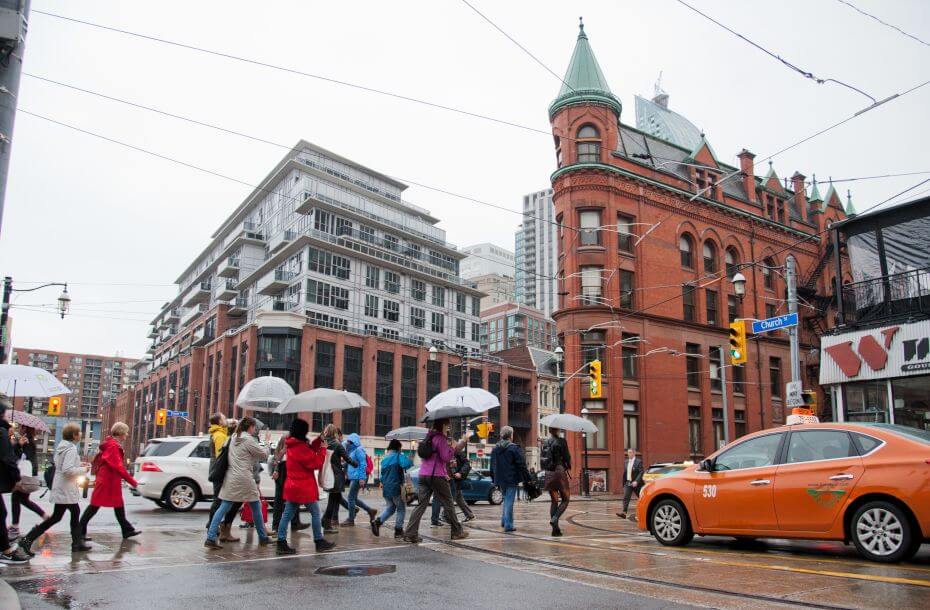 A group of people holding umbrellas cross the road on a busy intersection on a rainy day. The Flat Iron building can features prominently with its distinctive triangular shape.