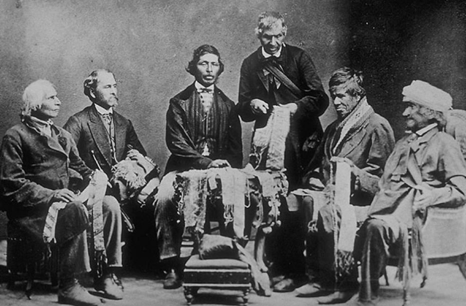 Black and white photo of five Indigenous men sit in a semi-circle on their laps they hold wampum belts that they are examining. One man is standing also holding a wampum belt. In front of the man in the centre is a table with multiple wampum belts on it. Bare wall in background.