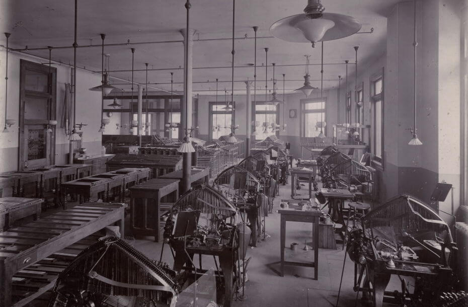 Black and white photo of a large room filled with machinery and equipment. Many windows are visible along the wall letting in daylight and lights hang from the ceiling above the machinery and work stations.