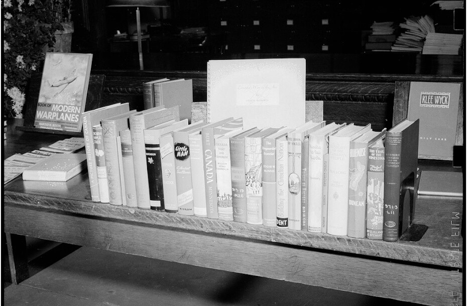 A black and white photograph of a row of books standing upright on a wooden table.