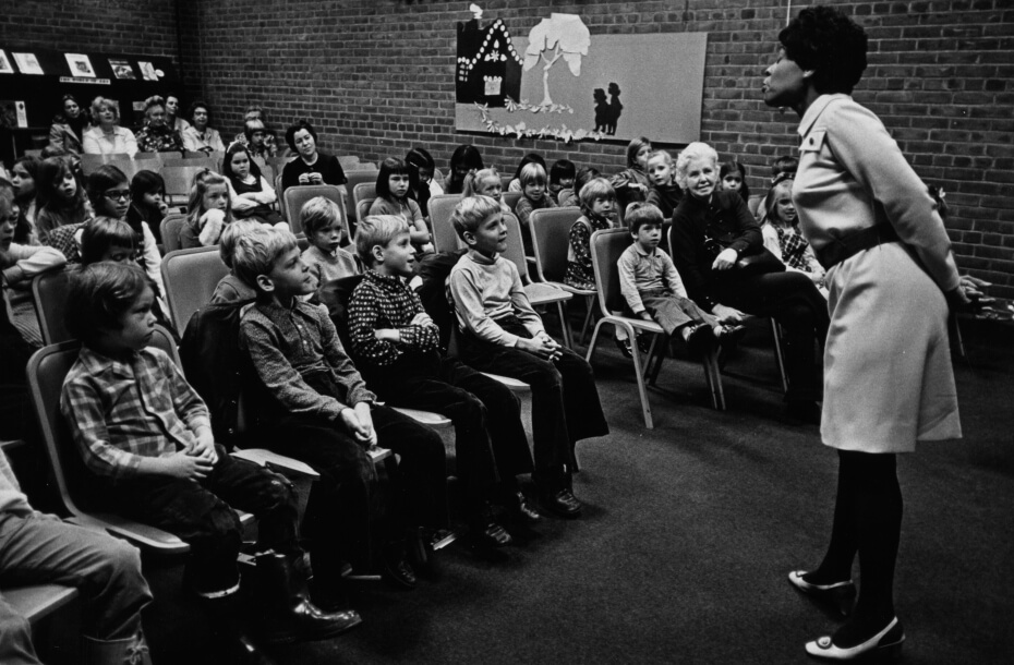 Black and white photo of a woman speaking a group of children sitting in chairs in a hall or classroom with brick walls. There is an illustration of a house on one wall.