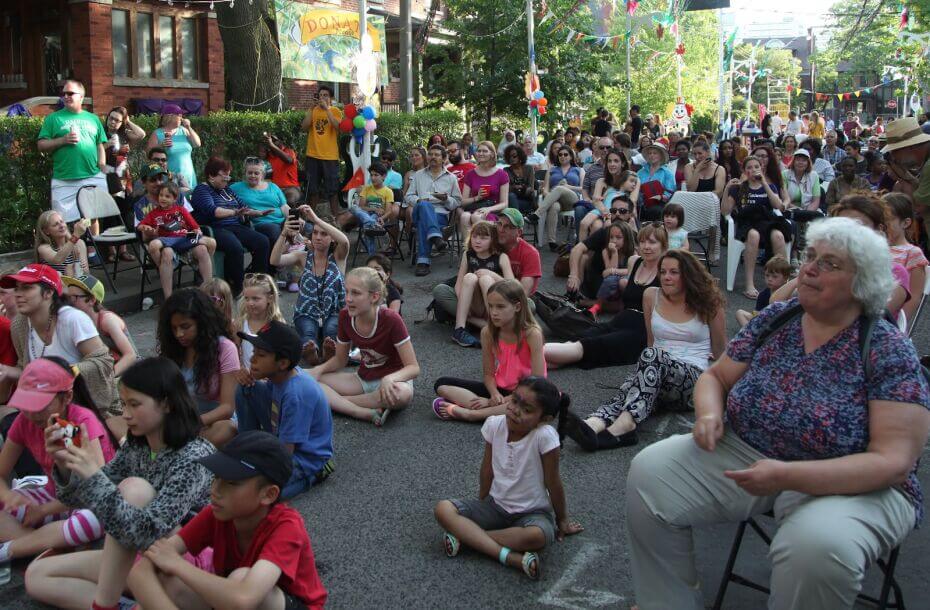 A large group on people sit in the middle of a paved road. In the front are several children sitting crossed-legged. There is also an elderly woman sitting on a chair. In the background there is a larger crowd of people also sitting on chairs. Two brick houses can be seen on one side of the street. On both sides of the street are tall, green trees with branches hanging above the crowd. The sun is coming through in the sky in between the two sides of the street.