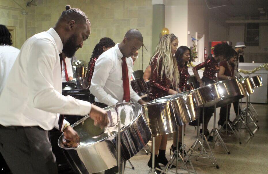 A group of individuals playing the steelpan. On the left of the photo there are two men in the first row wearing white shirts and burgundy ties. There are three women to the right of the photo wearing long sleeve, red sparkly dresses.