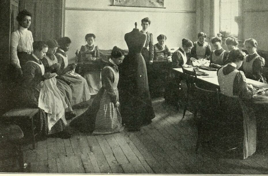 A black-and-white image of a group of women in dresses. Most of the women are sitting, either on a bench or at tables, and sewing. Two women are standing. In the center of the image is a sewing form with a dress on it.