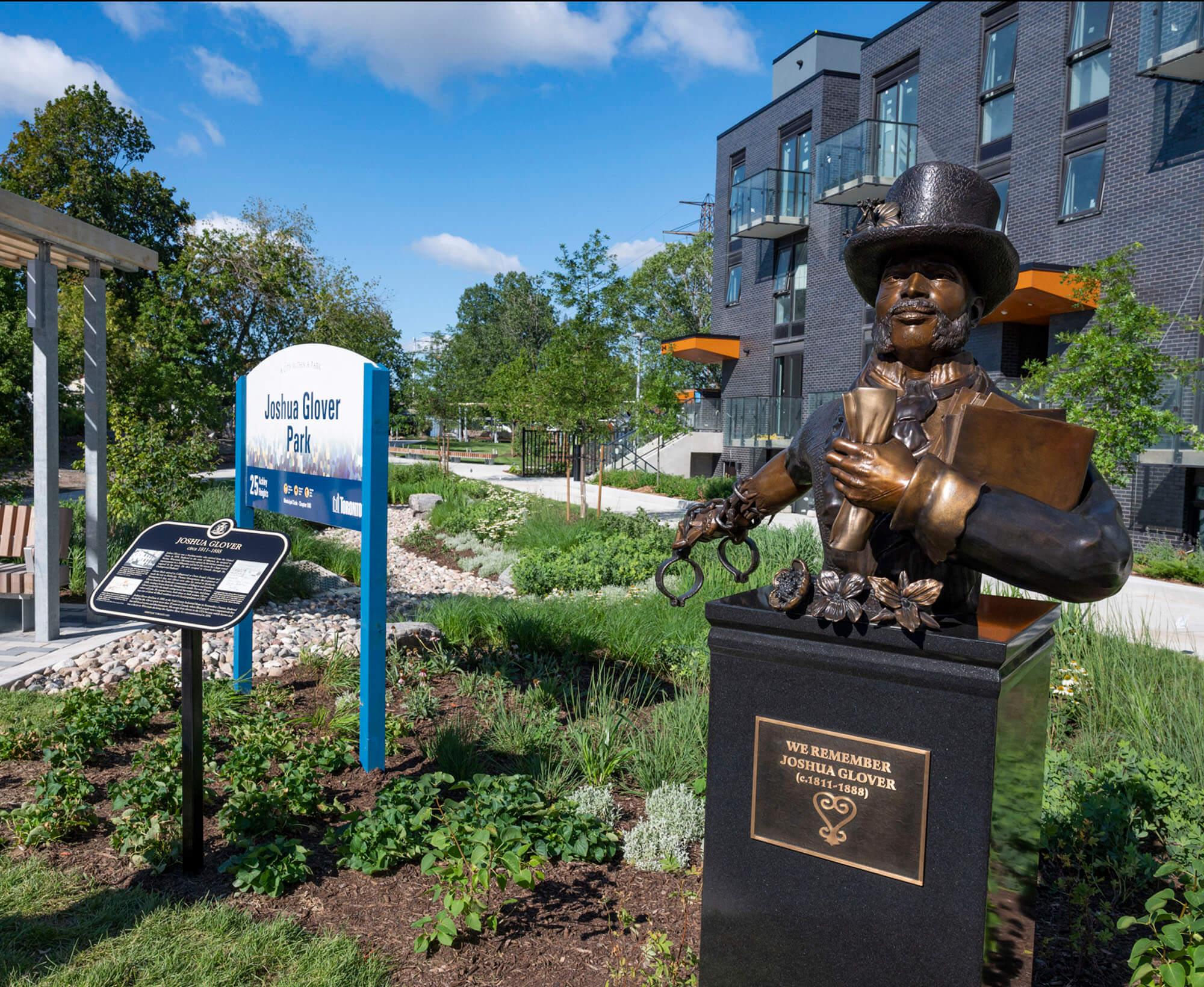 Image showing a park with bust of a man in a top hat on a pillar, a plaque on a post, and a sign reading Joshua Glover Park. Garden beds are visible with various plants and shrubs.