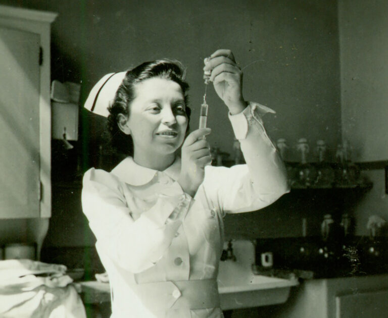 A woman in a nursing uniform holds up a syringe filled with fluid. She looks at the syringe. She stands in a medical room, filled with equipment.