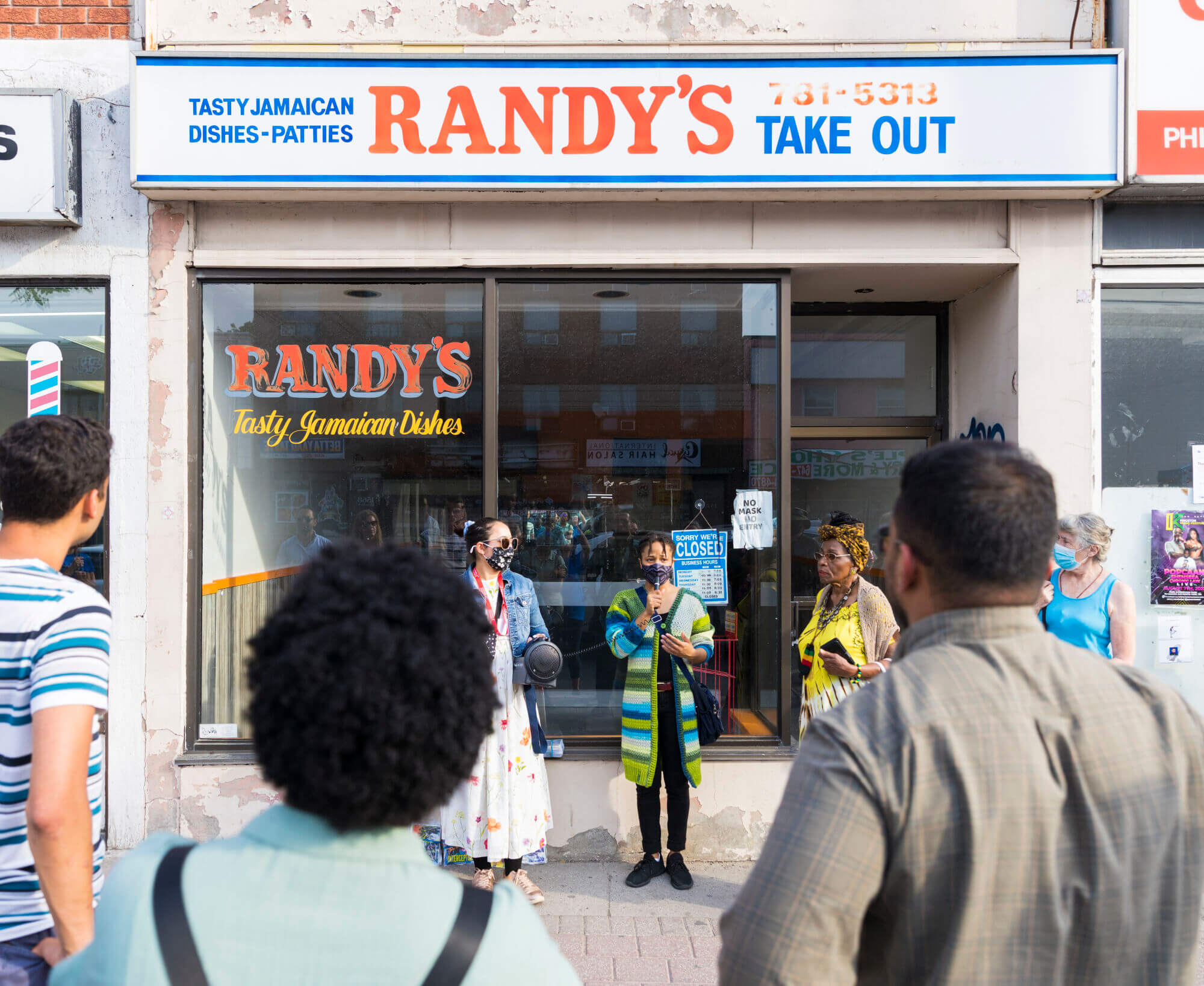 A woman is speaking to a group of people with a microphone and two people are standing beside her in front of a store with glass window panels at the storefront. The store sign above the windows reads " Randy's Take Out" in large blue and red lettering on a white background.