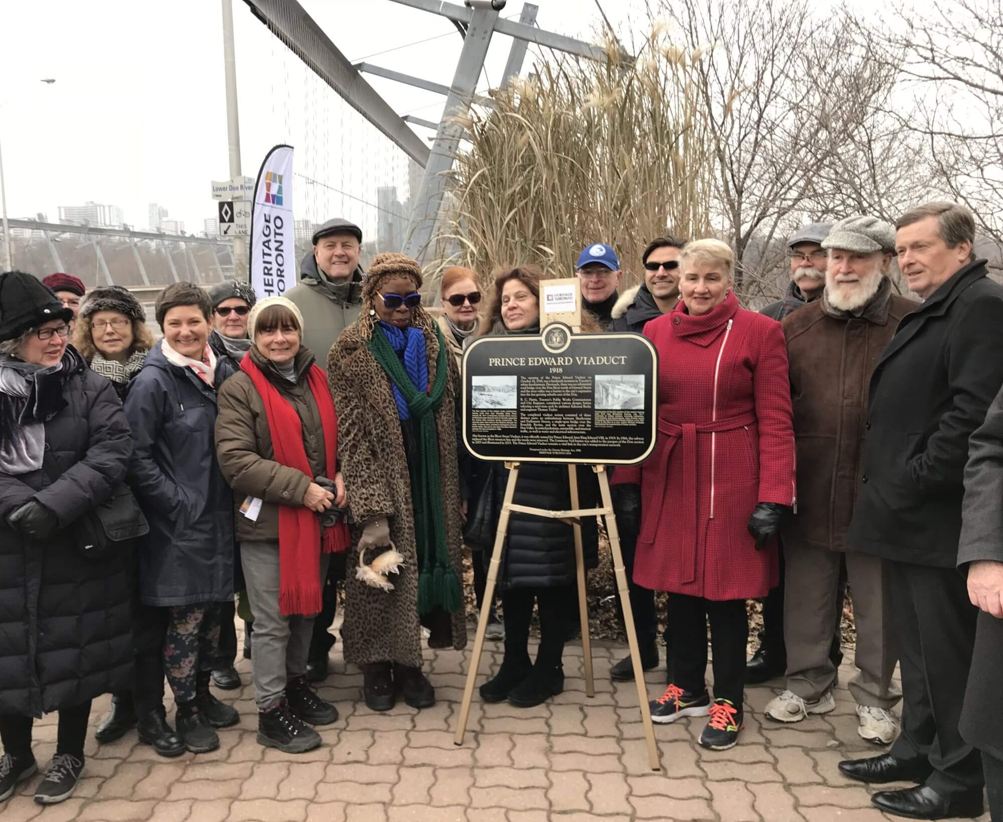 Colour photograph. A crowd of people stand behind the plaque, smiling for the photo. The Prince Edward Viaduct is in the background.