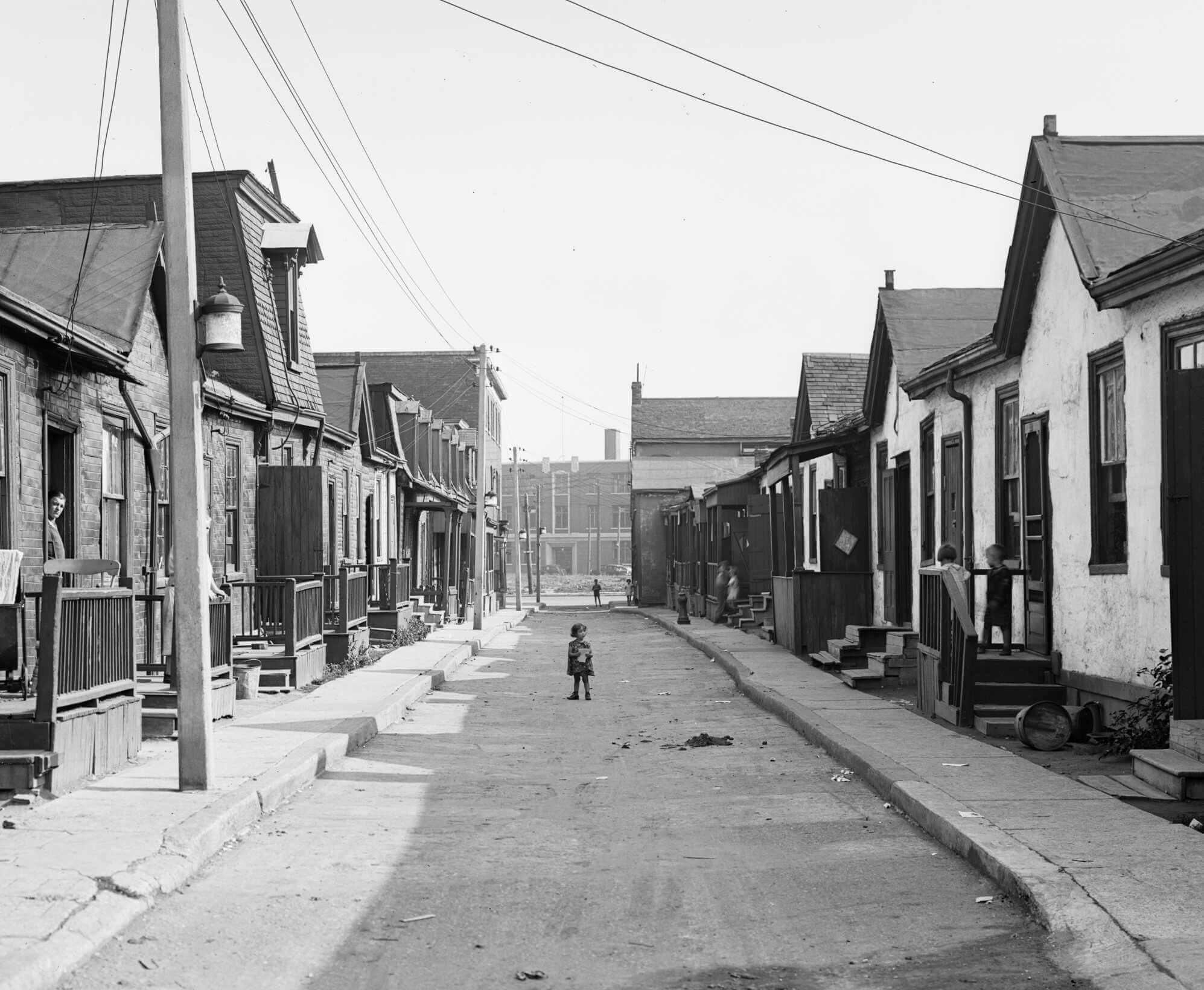 Black and white photograph looking down a desolate street. There are a few children scattered through the foreground and background playing games.