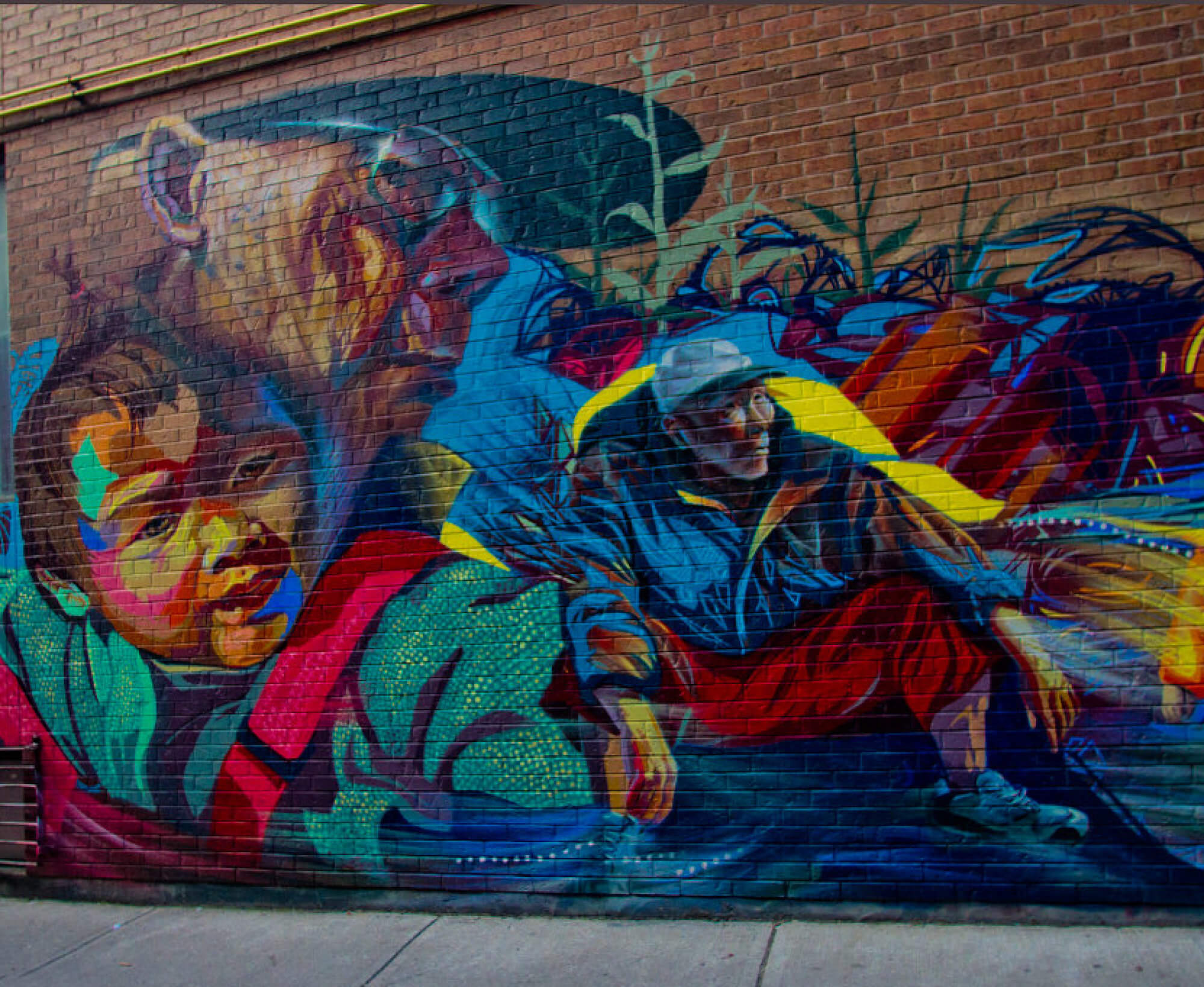 Colourful mural depicting a baby in a carrier in the foreground with two men wearing caps in the background, one is squatting. Vegetal motifs complete the vibrant mural.