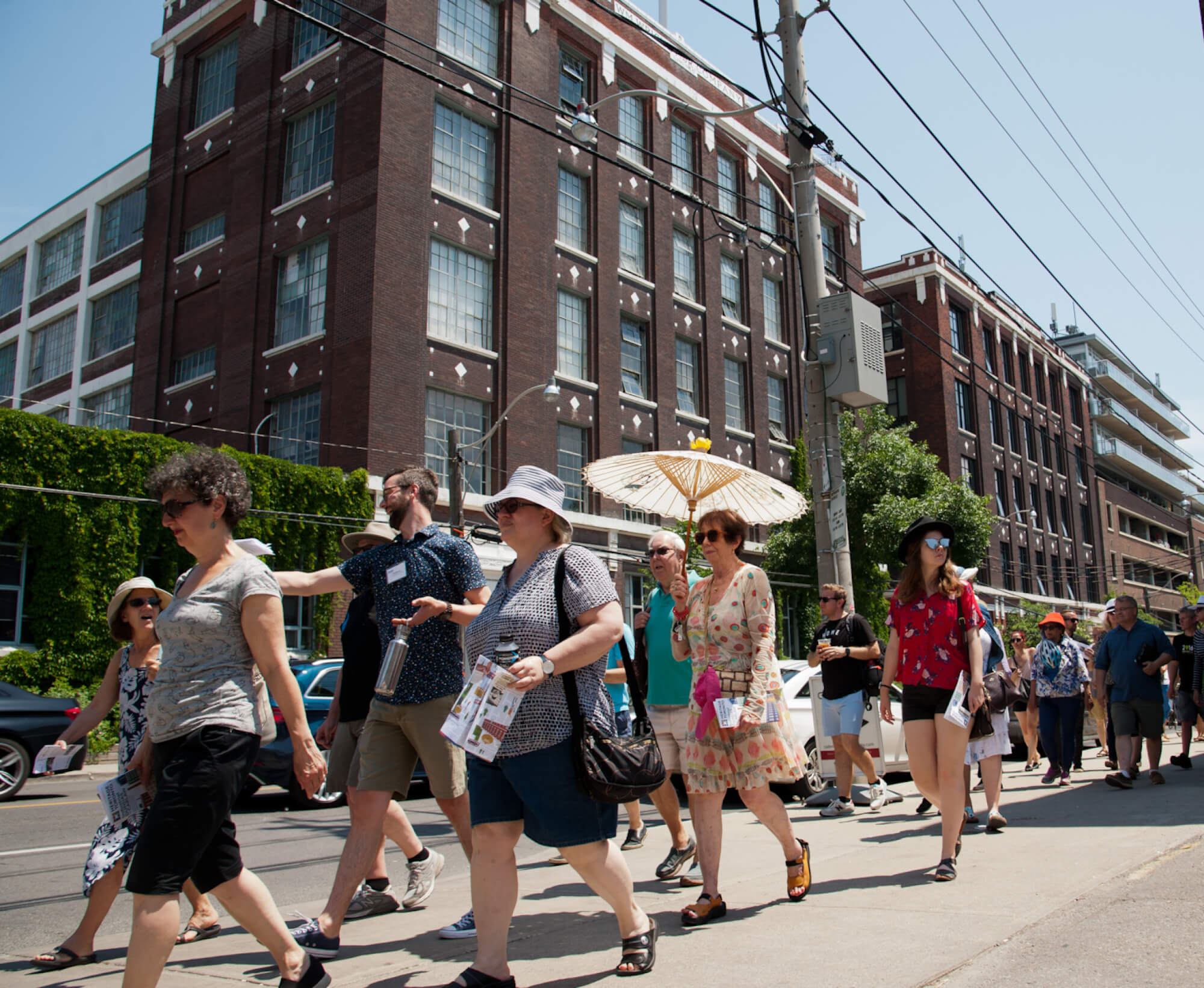 Image of people in summer clothing, including one woman holding an open Chinese umbrella, walk along a sidewalk in front of a five-storey brown brick building which fills the whole background of the photo.