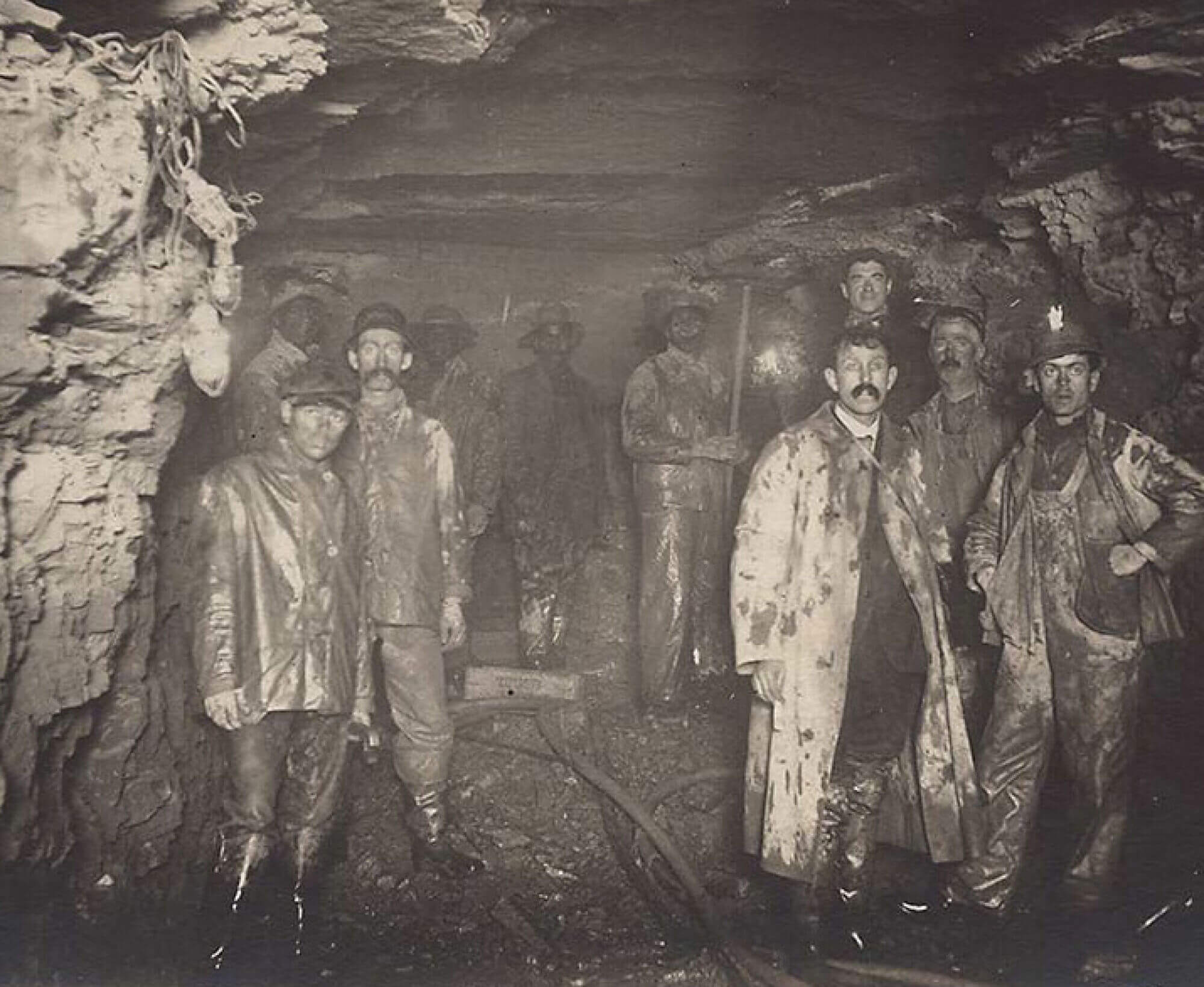 A black and white photograph of a group of men working in an underground tunnel. Their clothes are filled with mud and dirt.