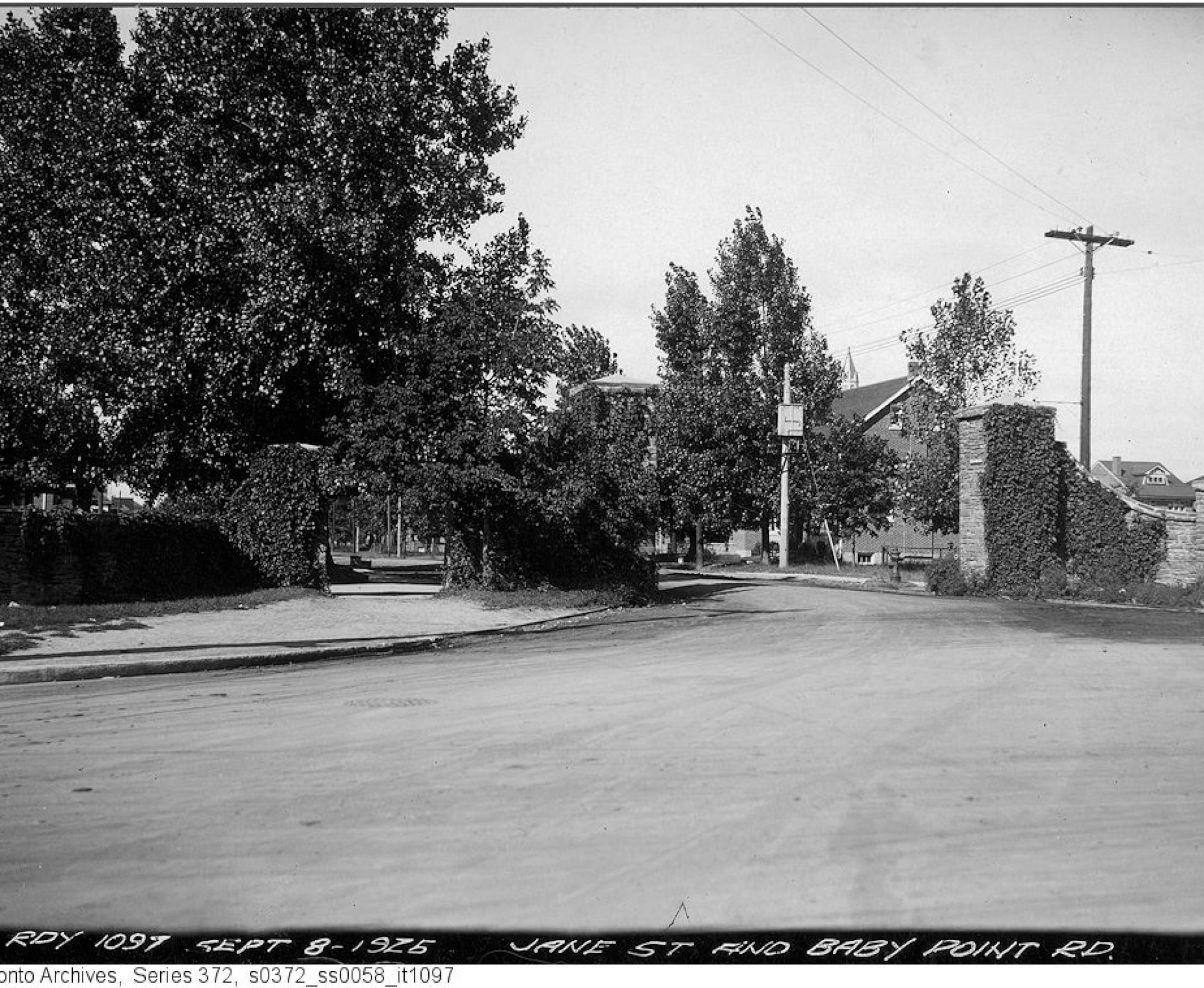 A black and white image of a three-way intersection. There are brick pillars on either side of the road covered in ivy. A large tree overhangs the road on the left side of the image.