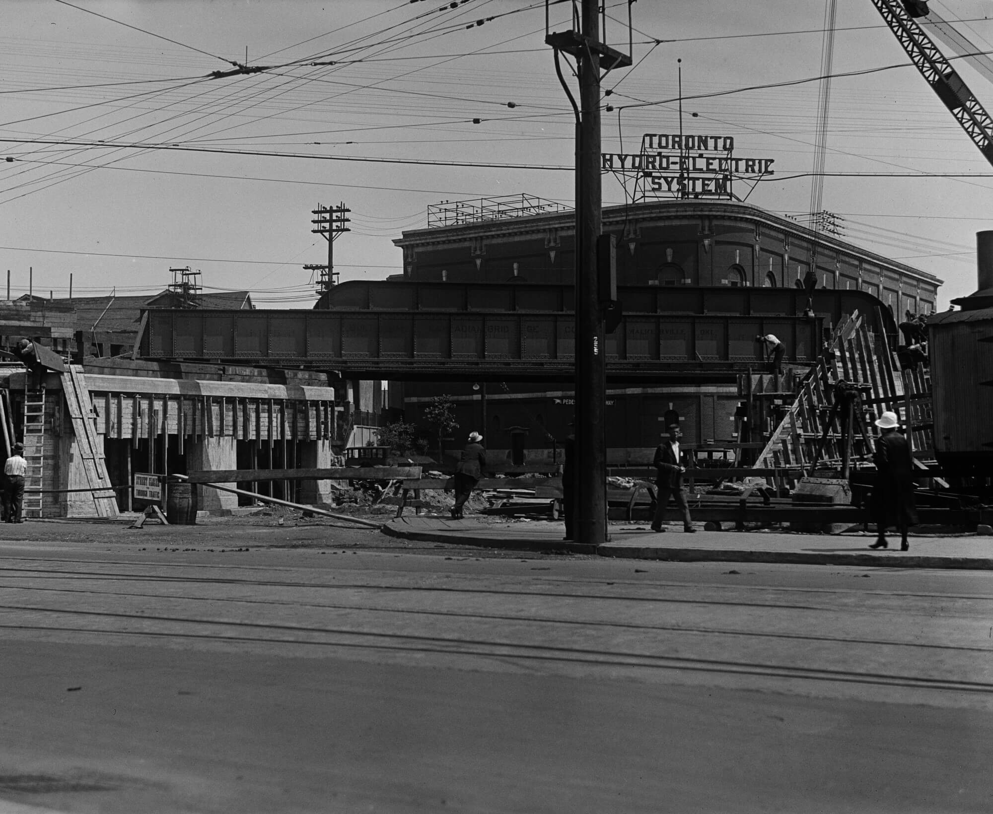 Black and white image of Carlaw Avenue from Gerrard Street East. Workers are building a rail bridge and a Toronto Hydro-Electric System substation is in the background.