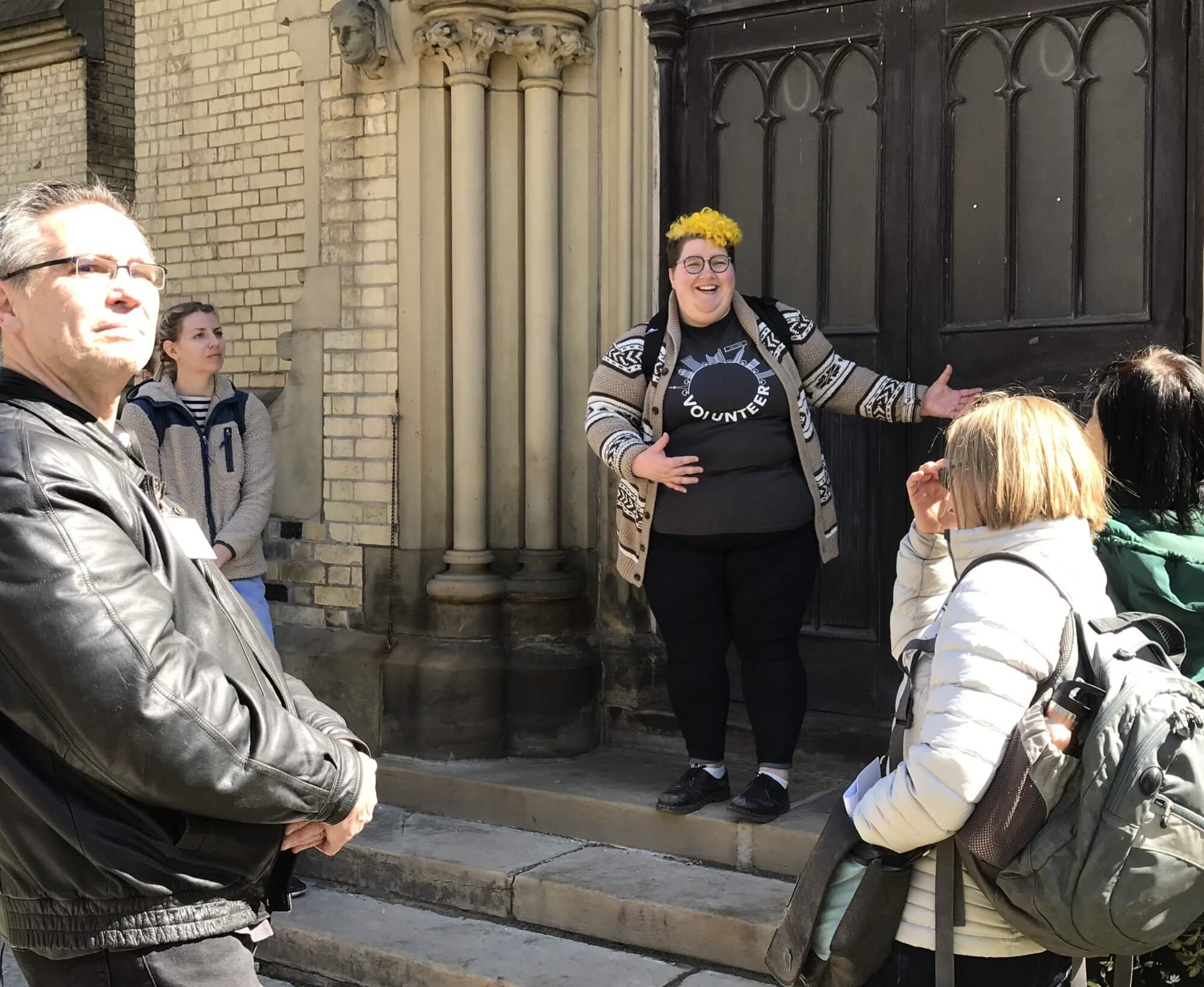 Image of young person standing on steps in front of side entrance of St James Cathedral. Participants of walking tour are gathered around them.