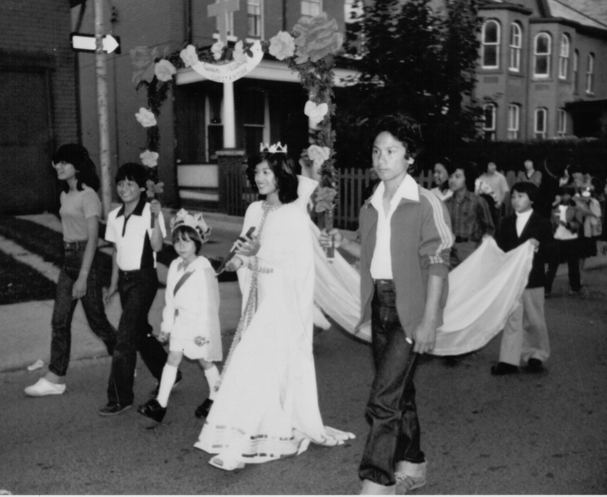 A black and white photograph of a Filipino Woman in a white dress walking as part of a procession for a Filipino ceremony, flanked by a man and three children, with a child carrying the train of her dress behind her.