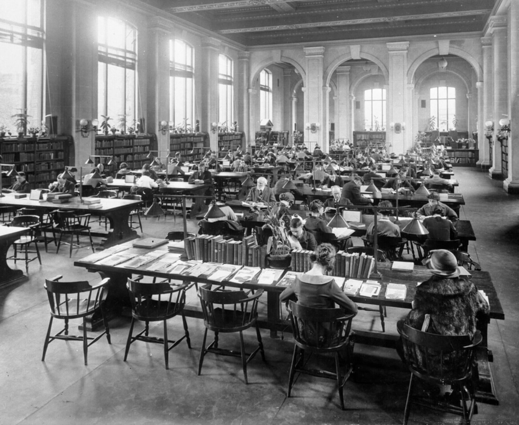 An archival photo shows people in a large room with high ceilings supported by tall arches and columns. There are wooden bookshelves lining the exterior walls while the interior has long wooden tables with people sitting and reading or examining documents.