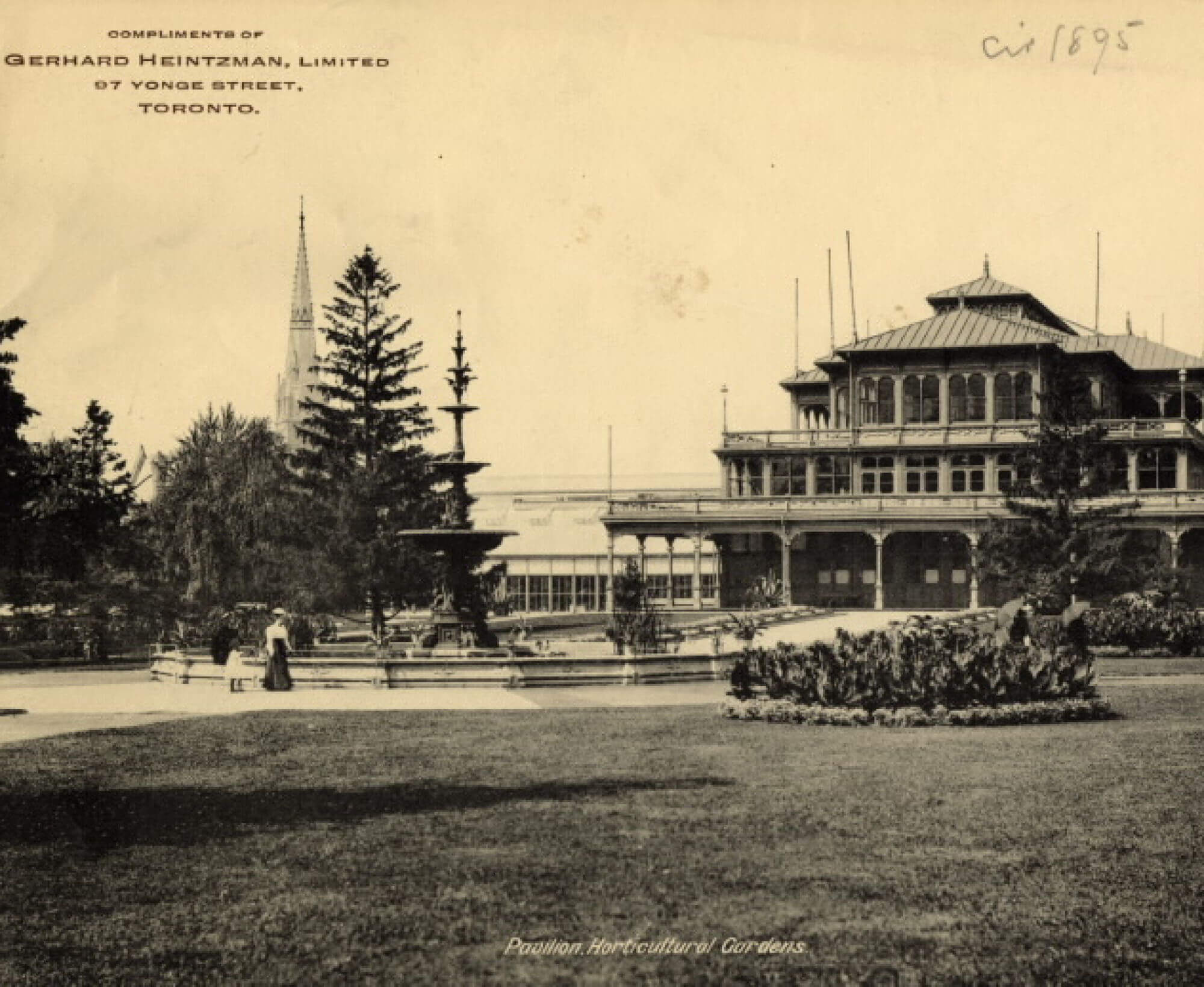 Sepia-toned photo of a multi-roofed building with a large sculptural fountain, pine trees, and large garden grounds.