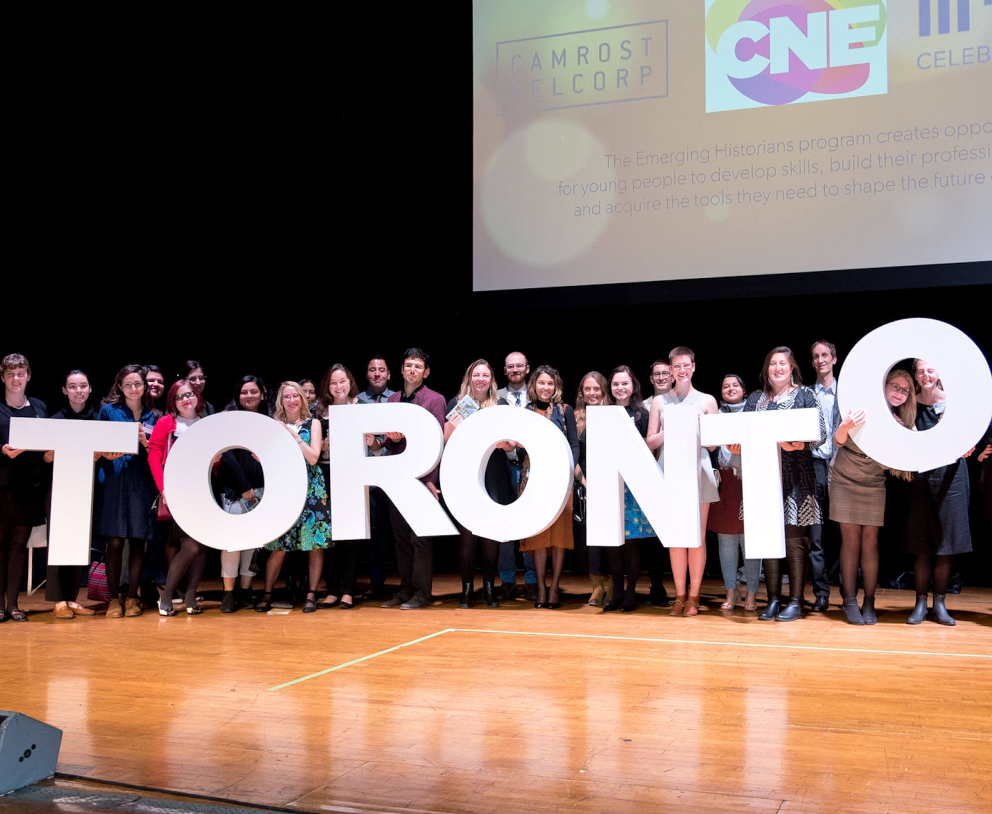 Image of 28 Emerging Historians, on stage at the 2019 Heritage Toronto Awards. They hold the letters for a mini TORONTO sign, and a projection screen appears above and behind them with logos of CNE, Tricon and Camrost Felcorp barely visible.