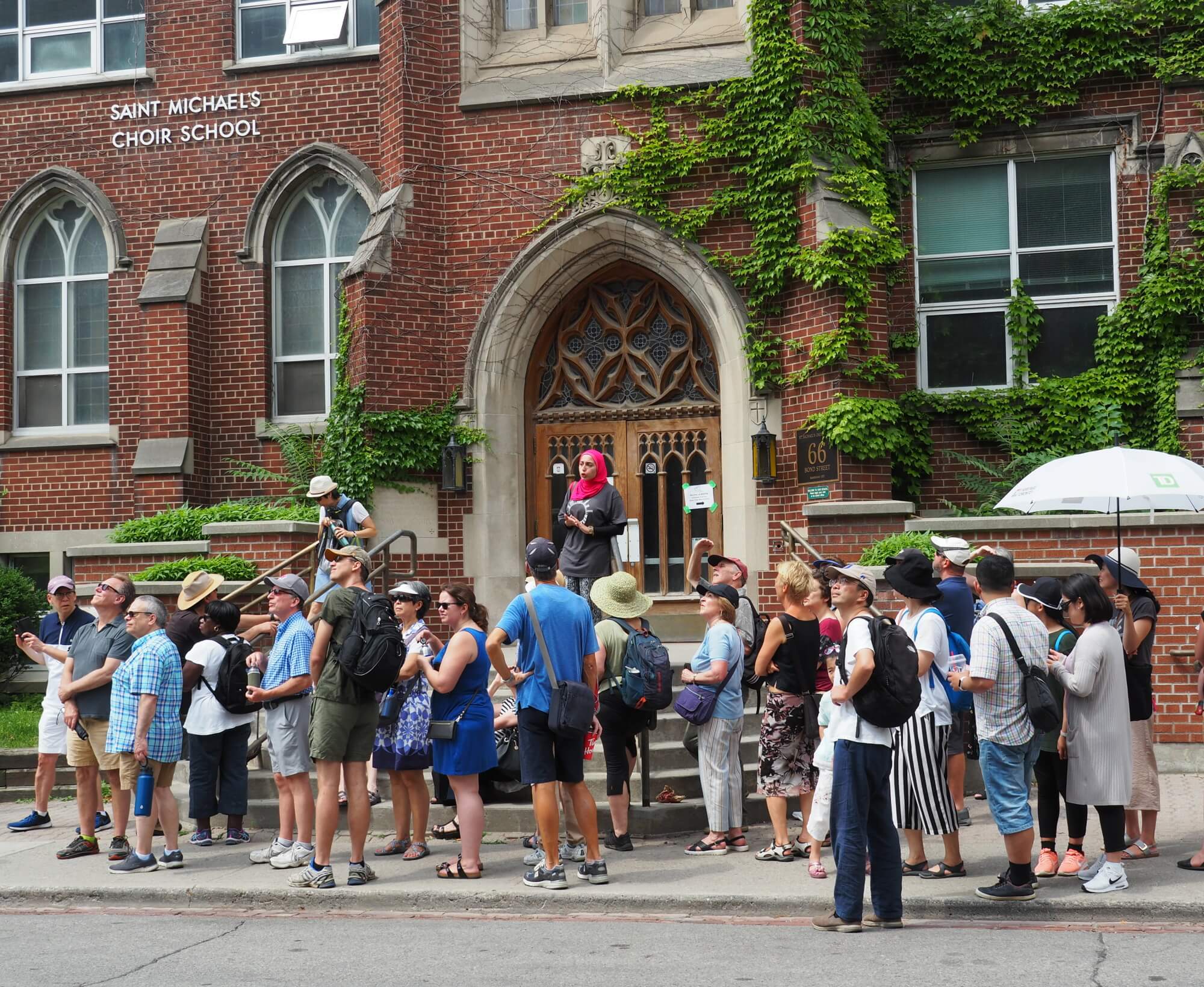 A large group of people in summer attire are standing along a sidewalk looking across road and are in front of a red brick building that features windows with pointed arches.