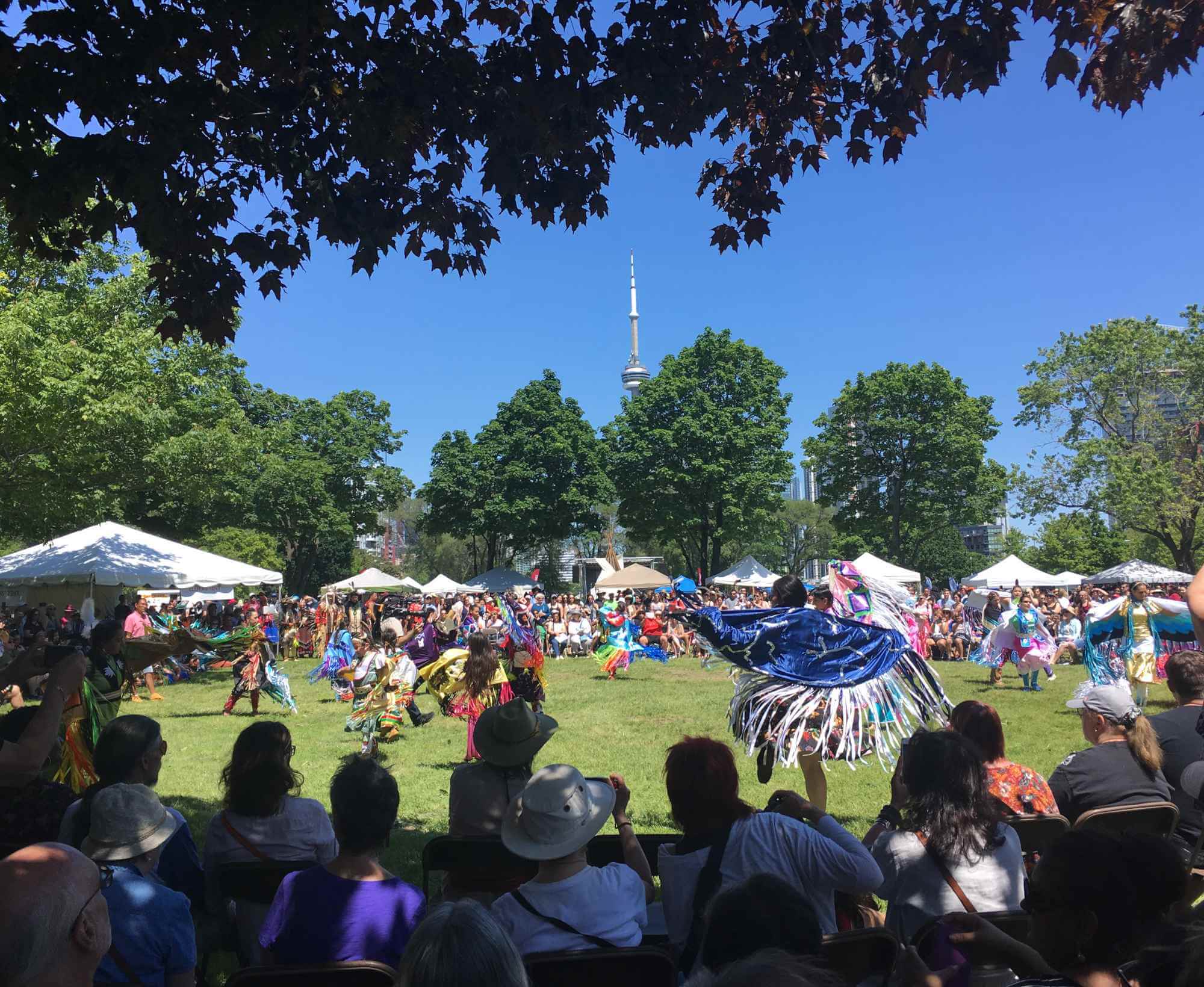 Colour photo of pow wow dancers in regalia dancing in a circle. Surrounding the circle are people watching the dancers. Beyond that are white vendor booths. In the background are trees and the top of the CN Tower can be seen in the distance.