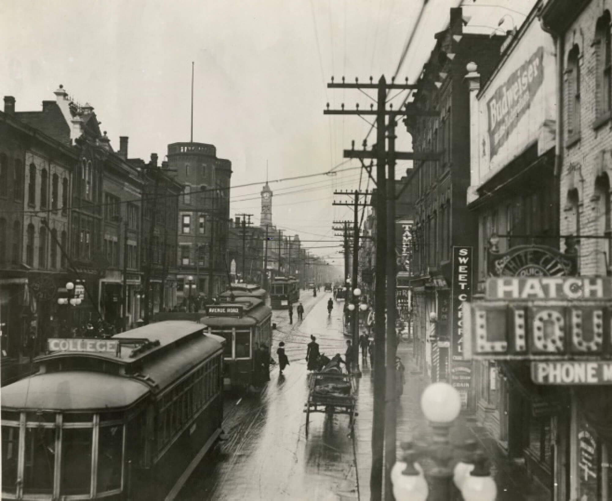 Black and white image of Yonge St from the vantage point of looking north from Granby St. Buildings of various architectural styles and electrical poles line the streets. Several of the buildings have electric signs hanging from the storefronts. Several streetcars are on the road, along with a horse-drawn carriage.