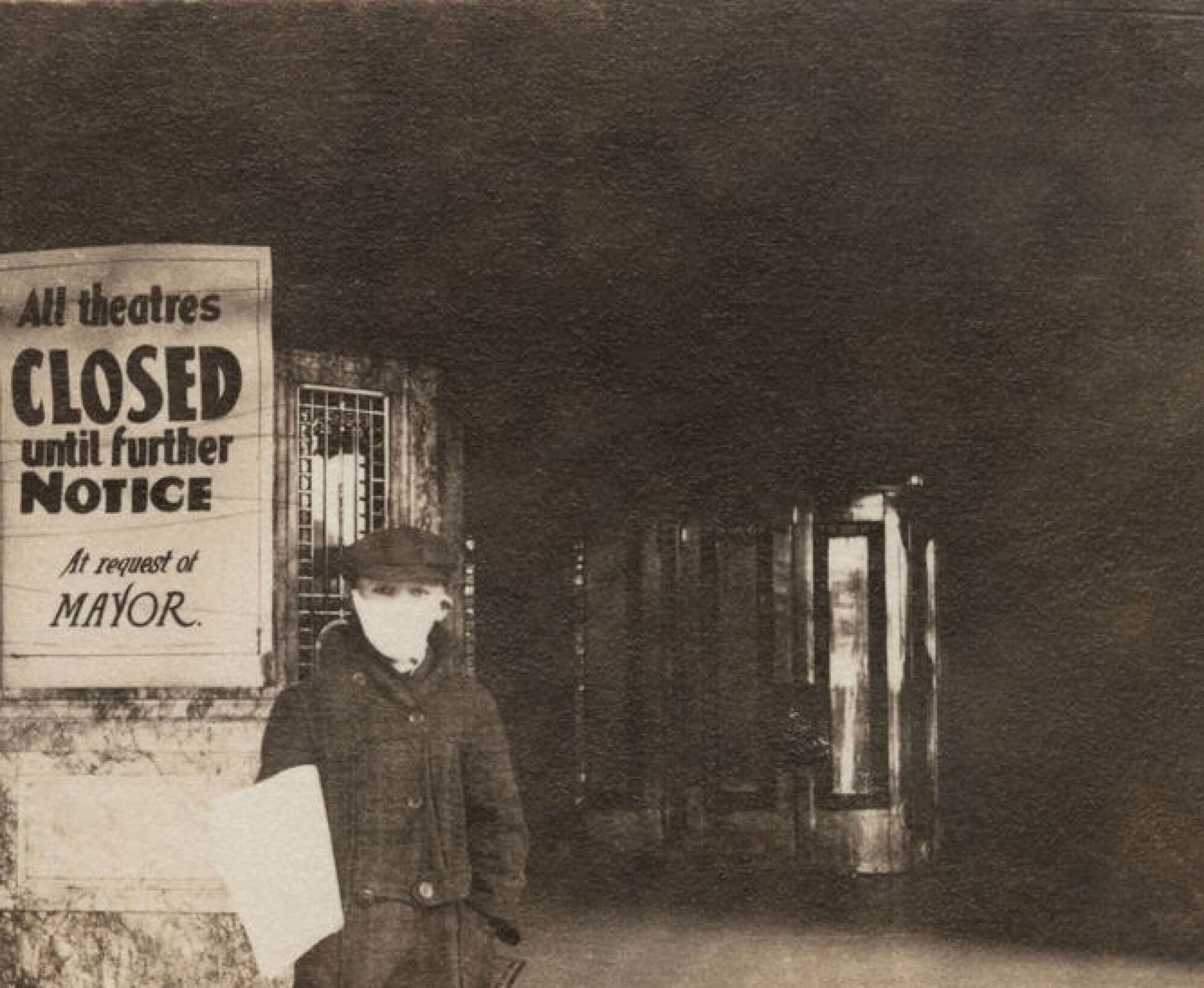 Sepia tone image of boy standing in front of a theatre entrance. He wears a mask. A sign posted on the box office reads - All theatres CLOSED until further notice. At request of Mayor