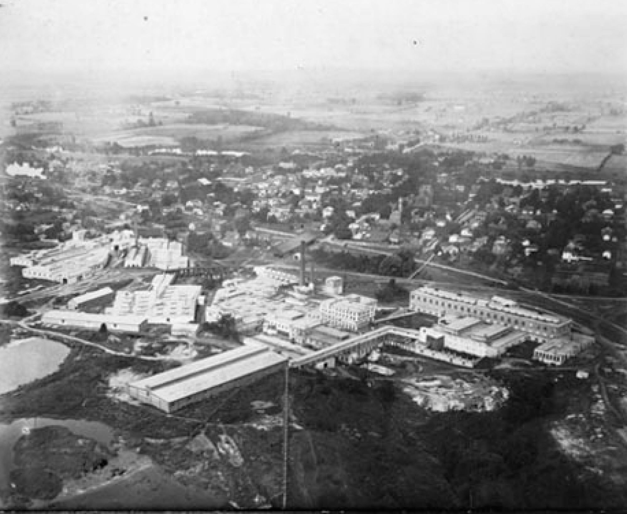 Aerial view showing a factory/industrial complex with several connecting buildings. Roadways are visible as are homes in a residential area in the background of the image.