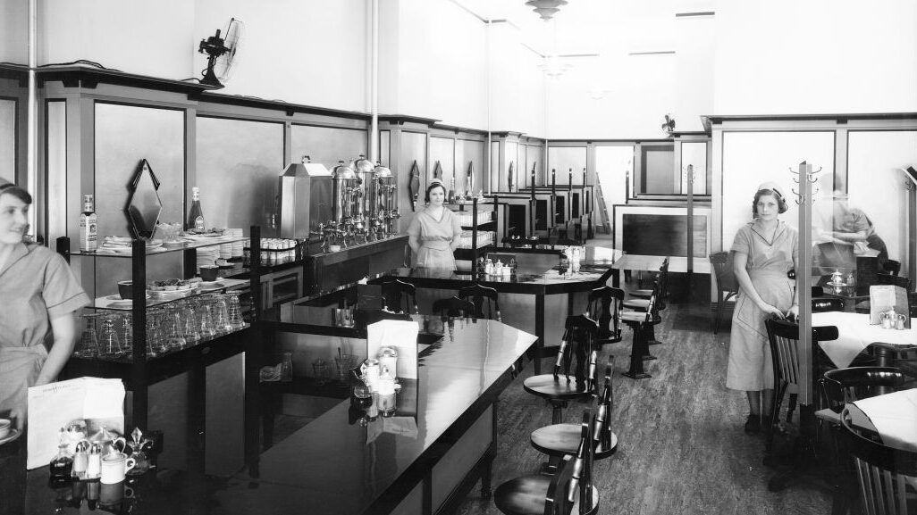 Black and white image of the interior of a restaurant, which features a counter and several tables. Three women in waitress uniforms are present: one at the far left, one in the middle, and one at the far right of the frame.