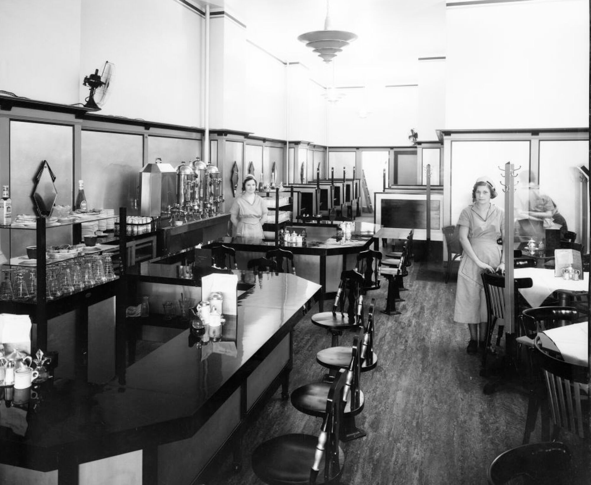 Black and white image of the interior of a restaurant, which features a counter and several tables. Three women in waitress uniforms are present: one at the far left, one in the middle, and one at the far right of the frame.